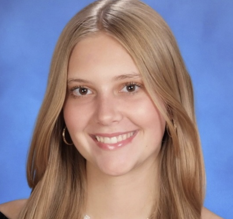 Young woman with long blonde hair, smiling, wearing hoop earrings, in front of a blue background.