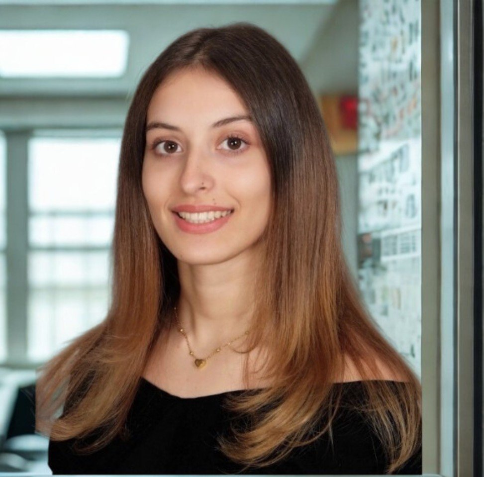 A young woman with long brown hair smiling in an indoor setting, with large windows and a bulletin board in the background.
