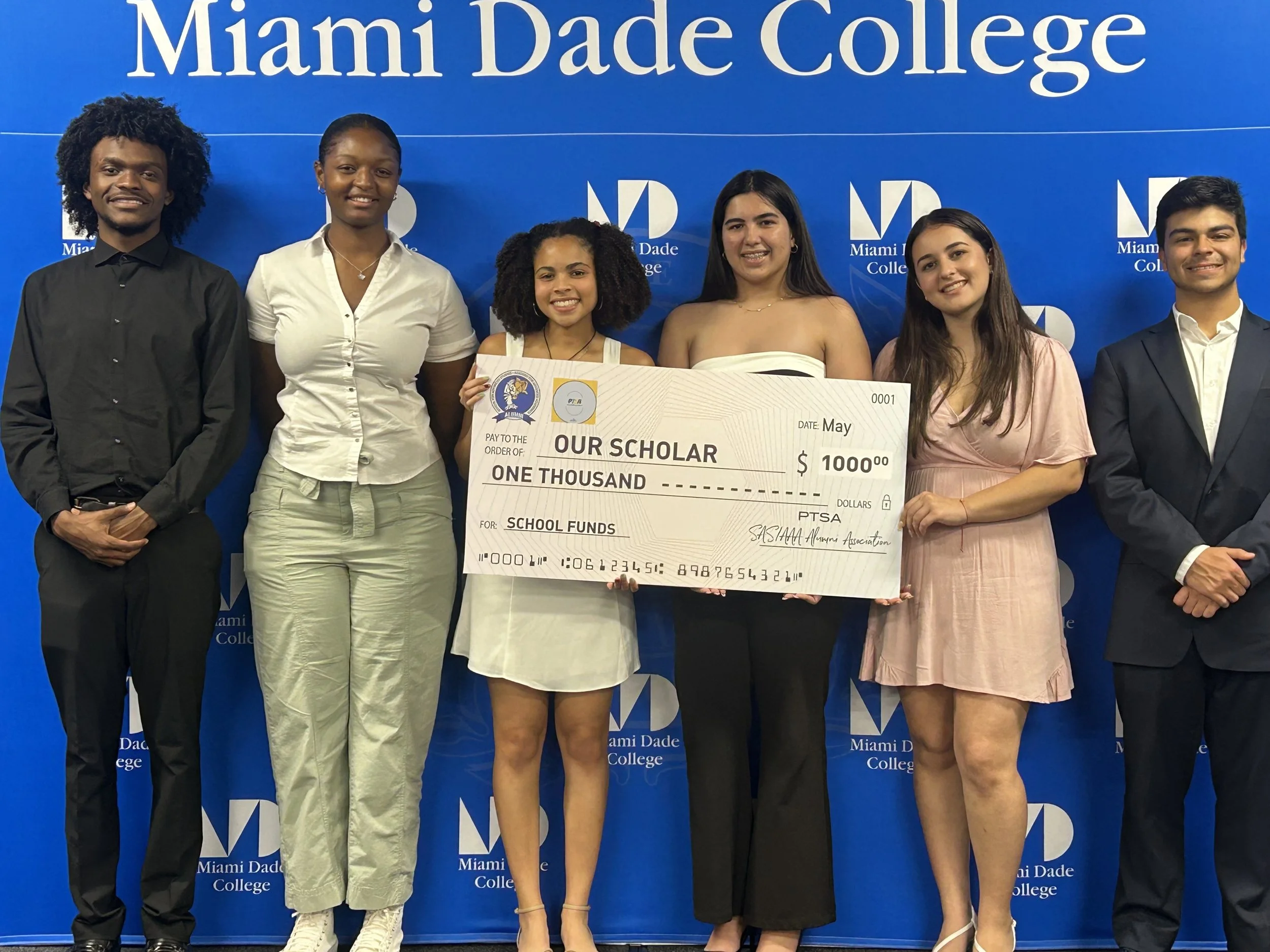 Group of six diverse young adults standing in front of a blue backdrop with the Miami Dade College logo, holding a large check for $1000 made out to 'Our Scholar' for school funds, celebrating an achievement.