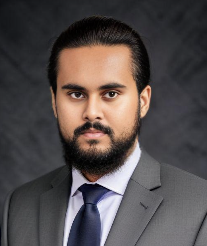 Portrait of a man with dark hair and beard, wearing a gray suit, white shirt, and navy tie, against a dark background.