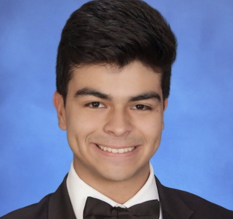 Smiling young woman with curly hair, wearing glasses, hoop earrings, and layered necklaces, in a well-lit indoor setting.