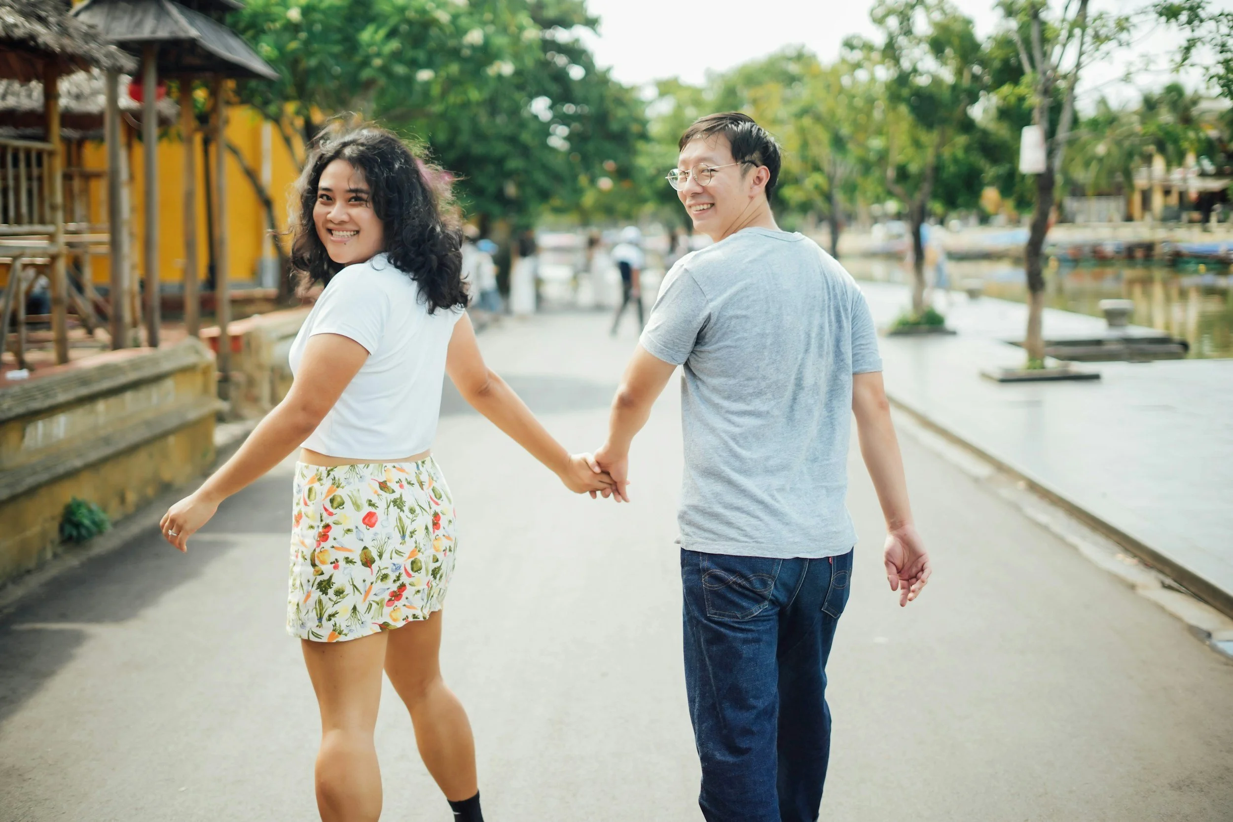 A smiling woman and man holding hands while walking outdoors on a sunny day near a waterway with trees and buildings in the background.