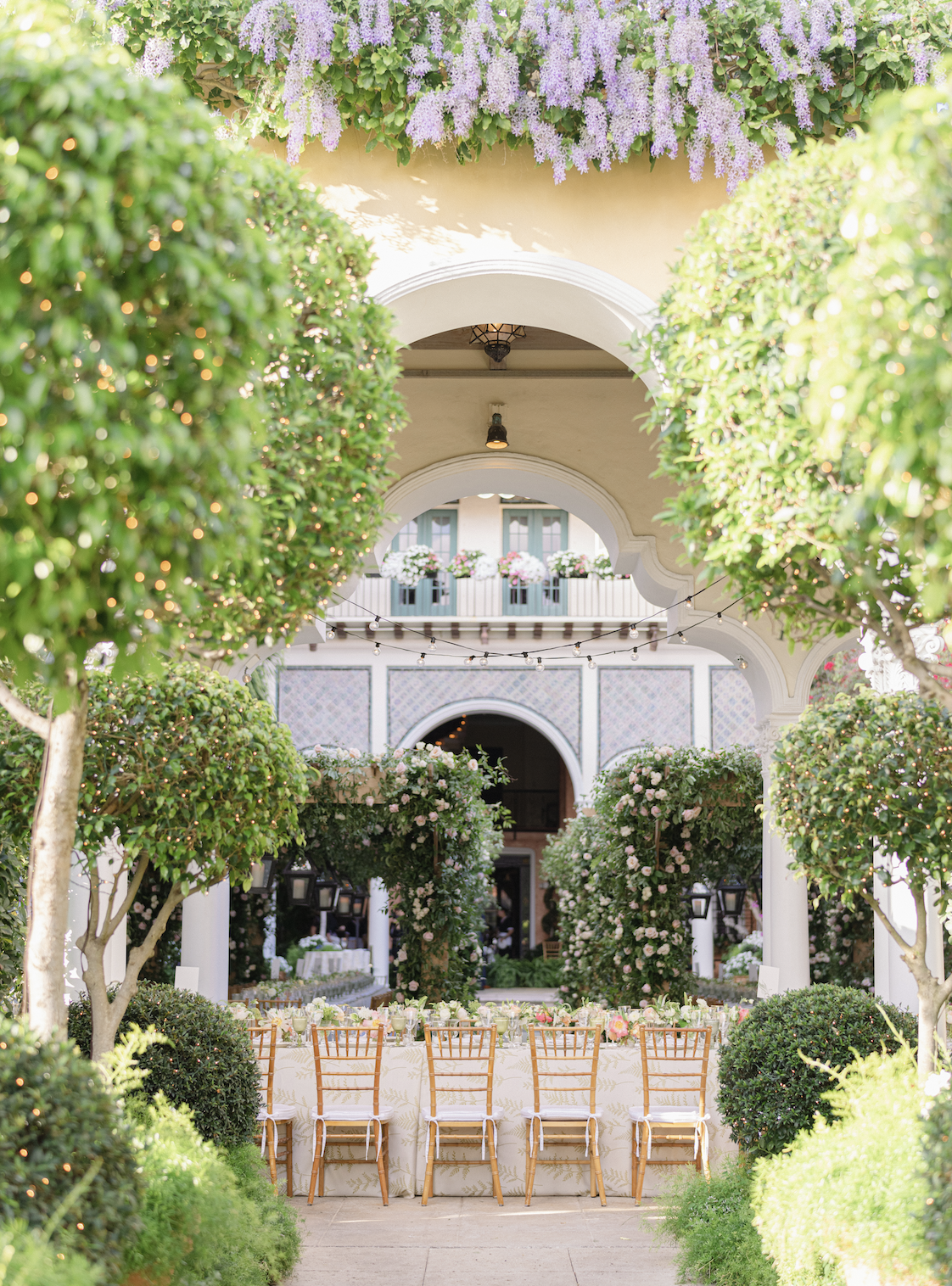 A decorated outdoor garden area with a long table set for a gathering, surrounded by lush greenery, flowering trees, hanging lights, and an arched structure with potted flowers above.