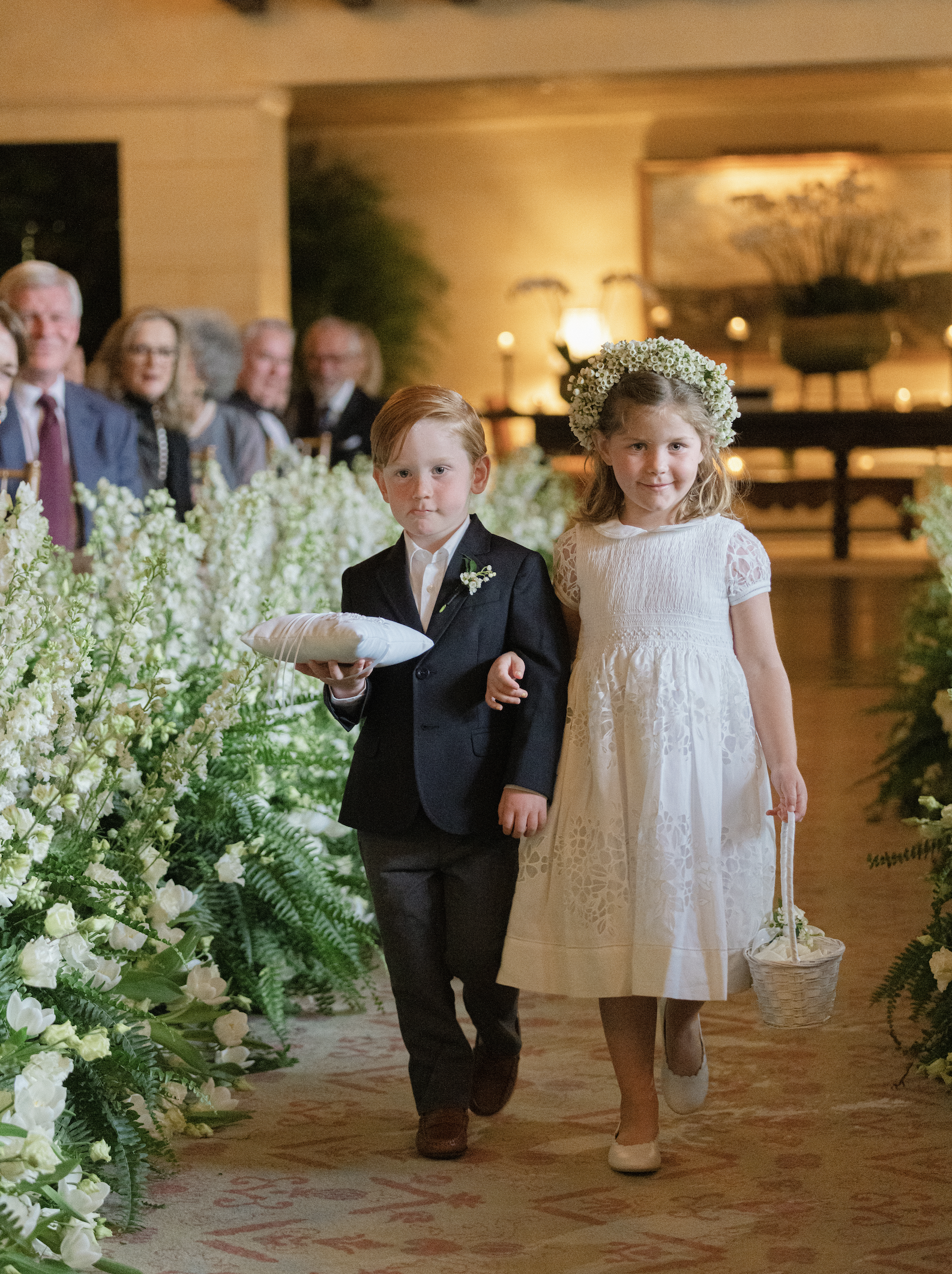 A young boy and girl walking down an aisle during a wedding ceremony. The boy is dressed in a suit and holding a pillow, and the girl is in a white dress with a floral crown and basket. Guests are seated in the background, with warm lighting and flor