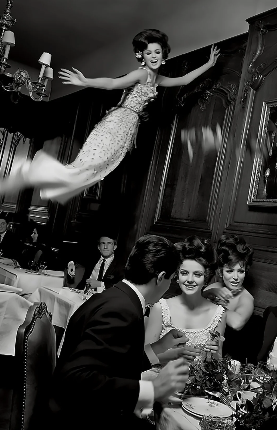 A black and white photograph of a woman floating or flying in a formal dining room, wearing an elegant dress, with a group of people seated at a table below, engaged in conversation.