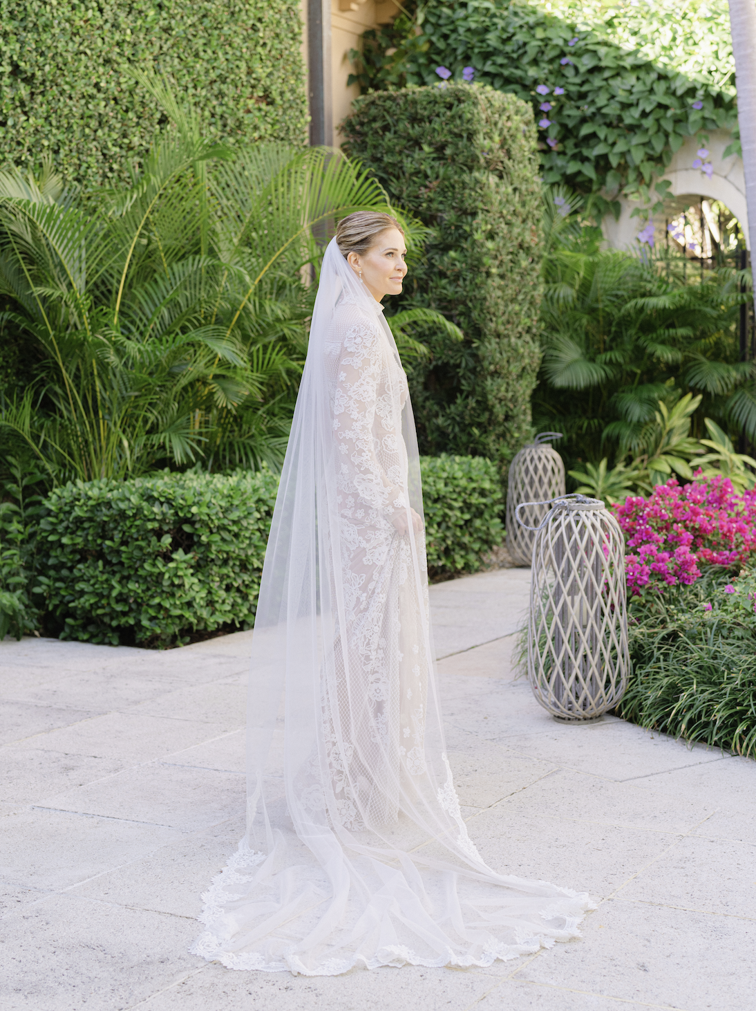 Bride in a white lace wedding dress and veil standing outdoors in a garden with lush green plants and pink flowers.