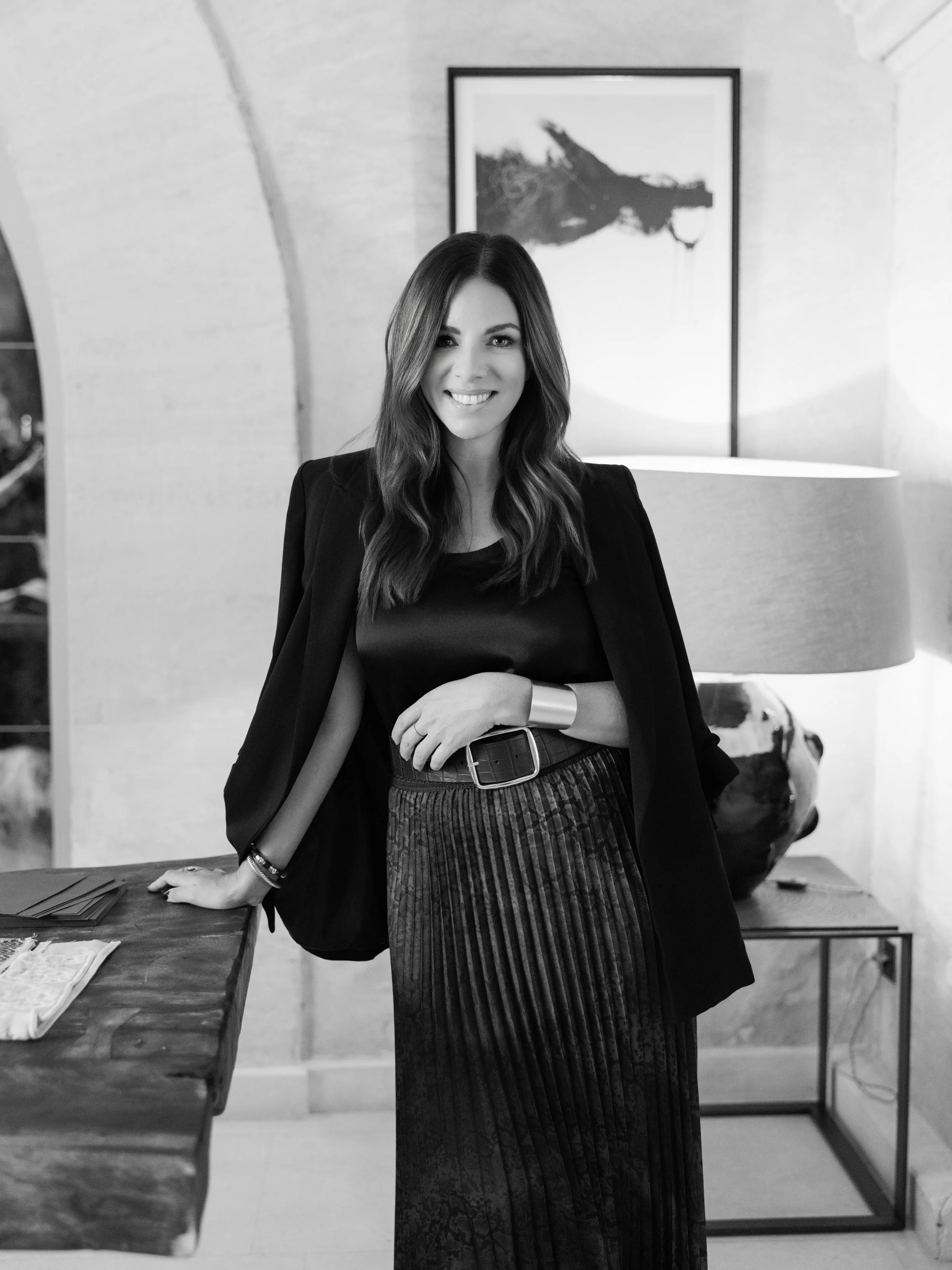A smiling woman with long wavy hair wearing a black blazer and a pleated skirt, standing indoors next to a wooden table with fabric samples, in front of abstract artwork and a lamp.