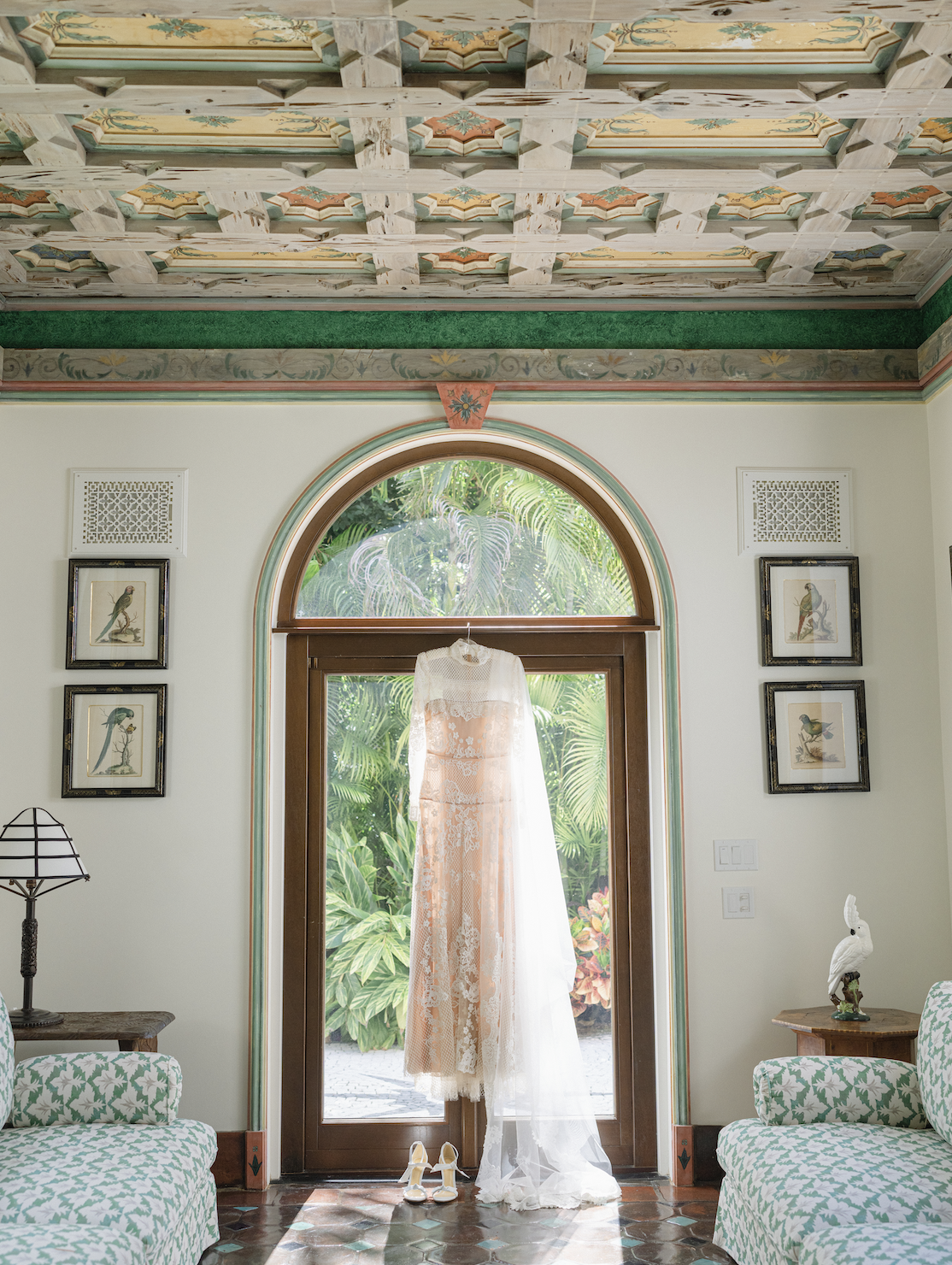 A wedding dress with lace details hanging in front of a glass door with greenery outside, flanked by framed bird illustrations and surrounded by patterned furniture and decor in a room with a decorated ceiling.