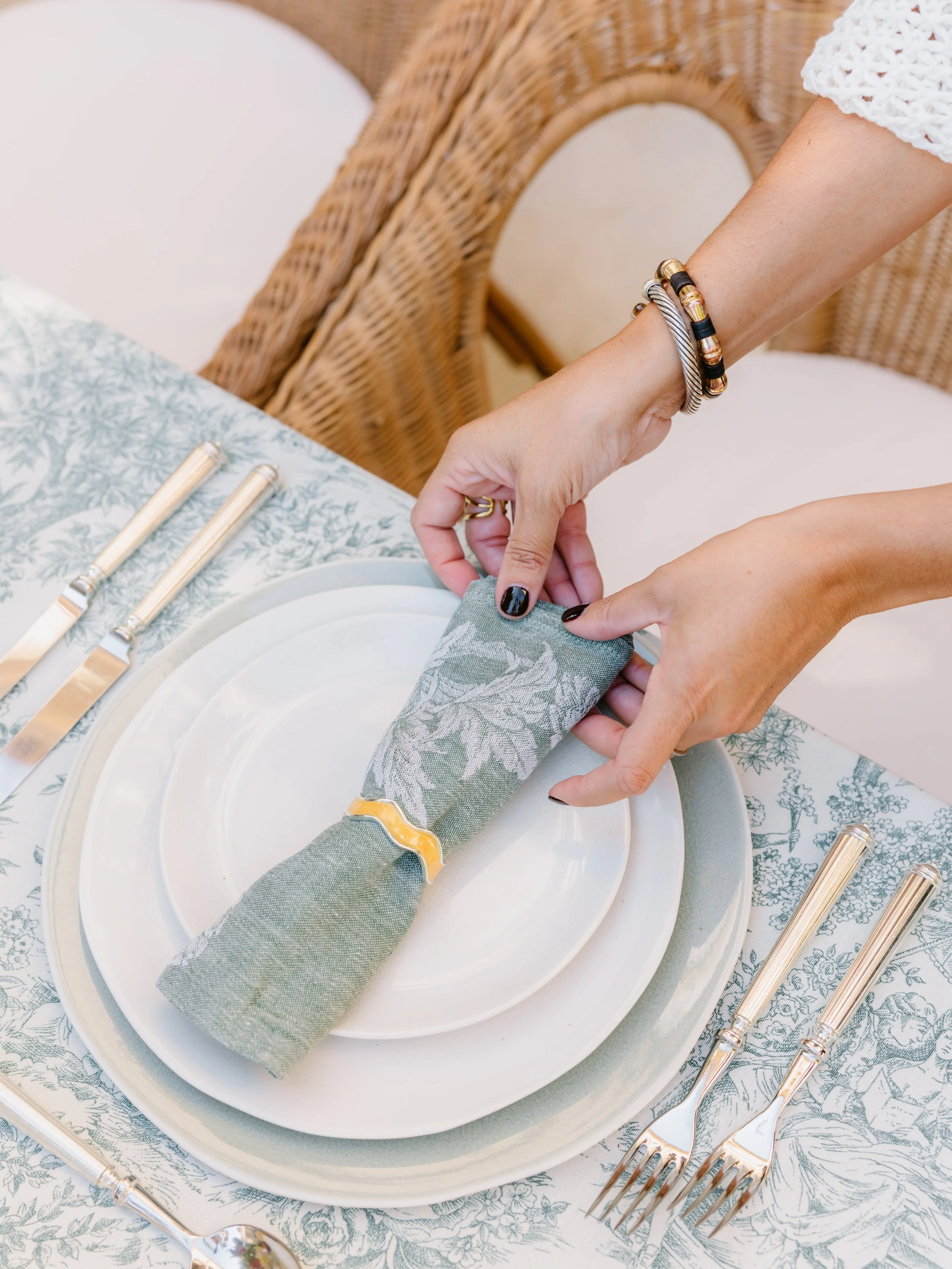 Person setting a table with a green linen napkin, white plates, and silverware, with wicker chairs in the background.