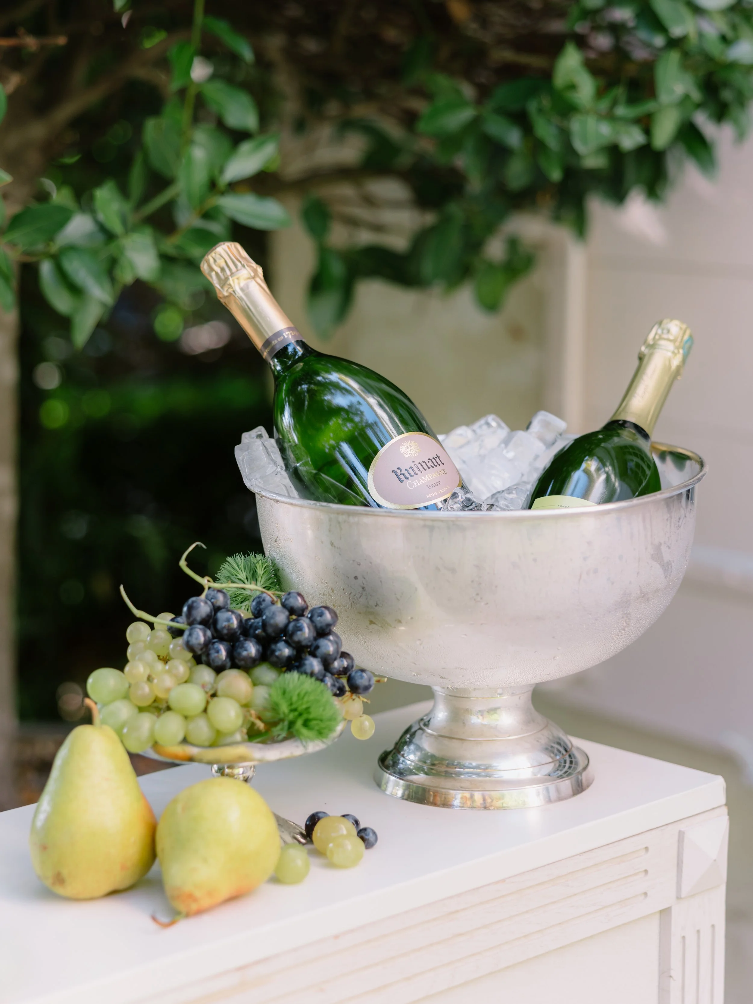 Two bottles of champagne in a silver ice bowl on a white surface with fresh grapes and pears in front, outdoors with greenery background.