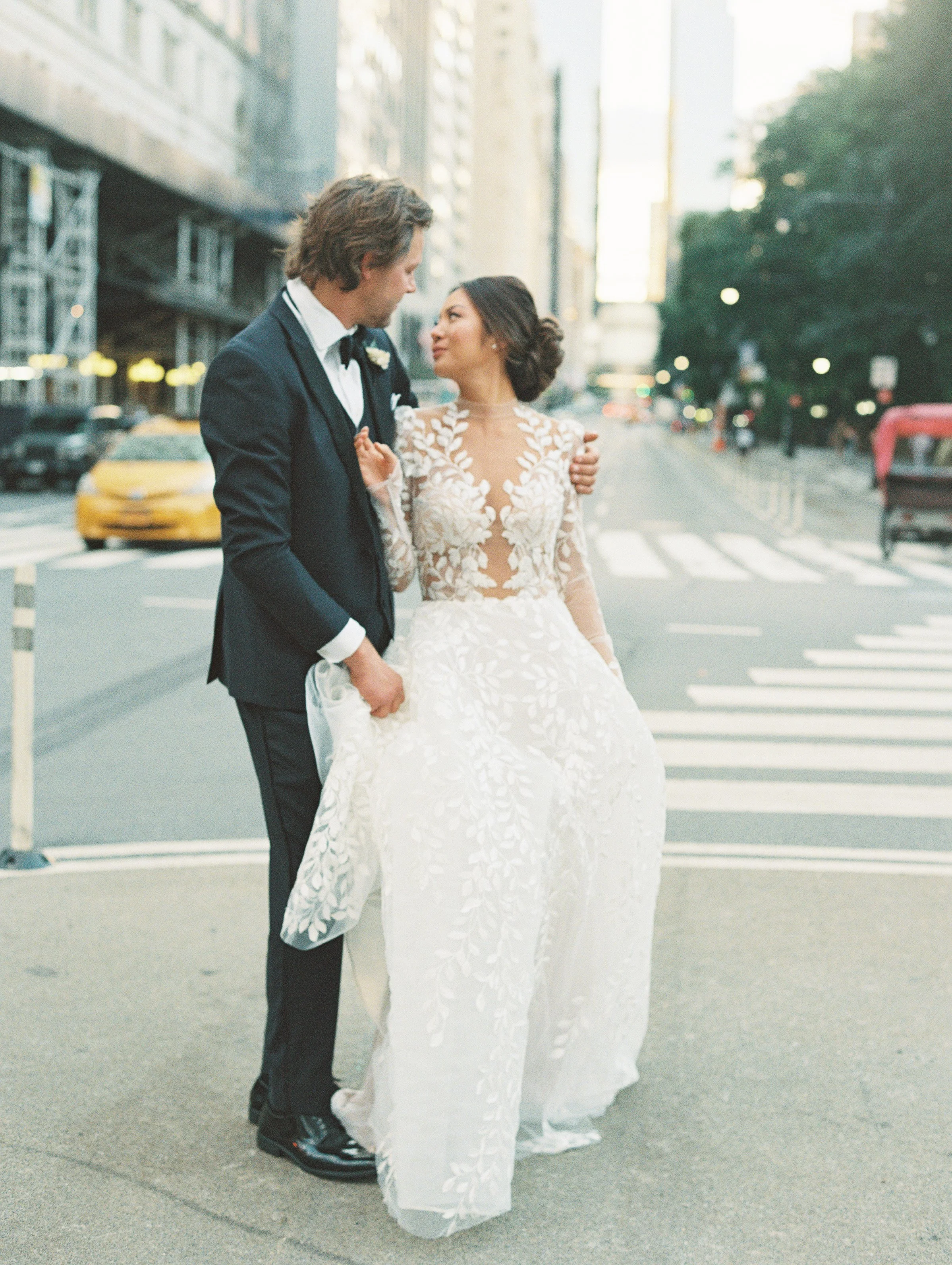 A bride and groom in wedding attire sharing a moment on a city street at sunset.