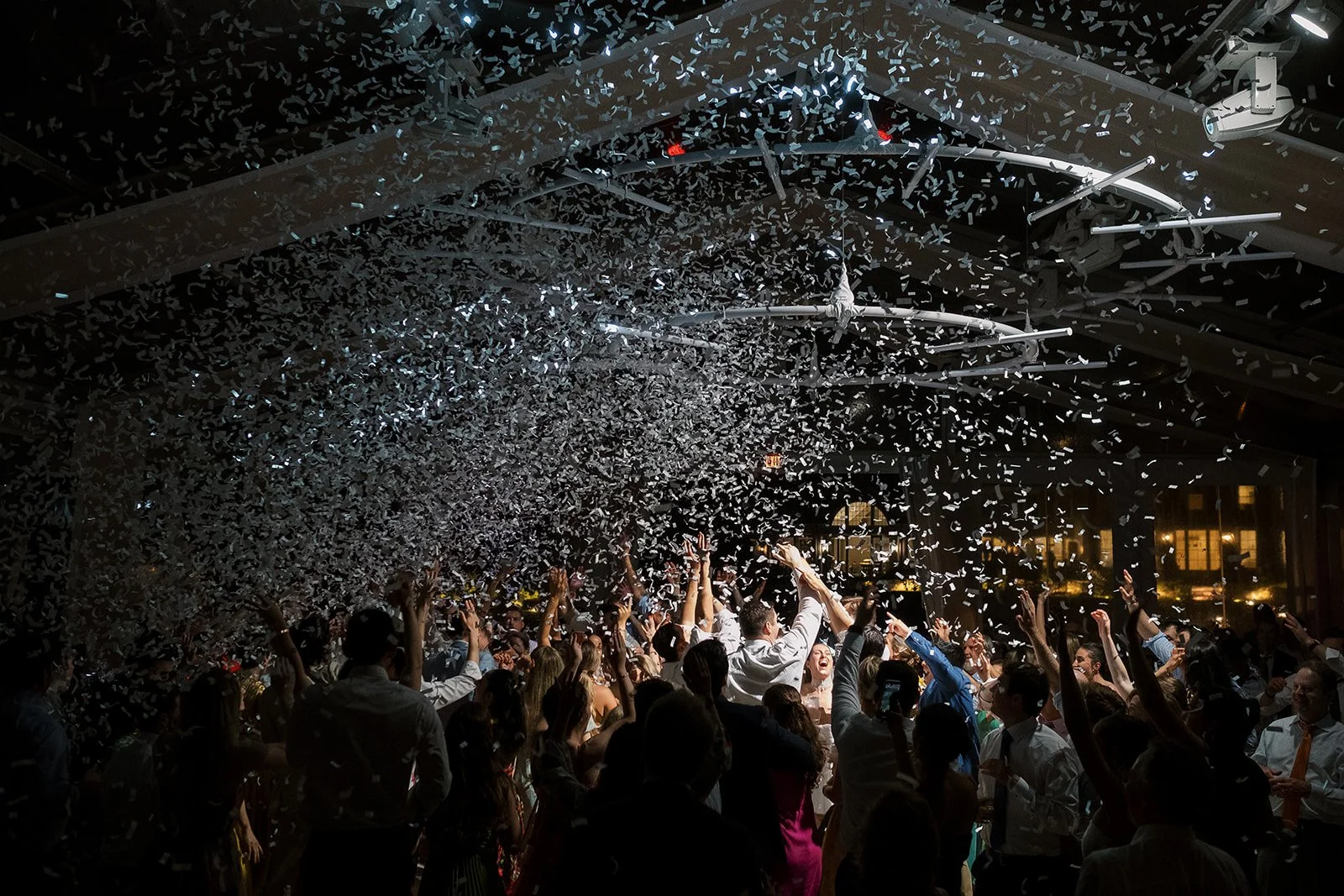 Crowd of people celebrating with confetti falling inside a decorated event space at night.