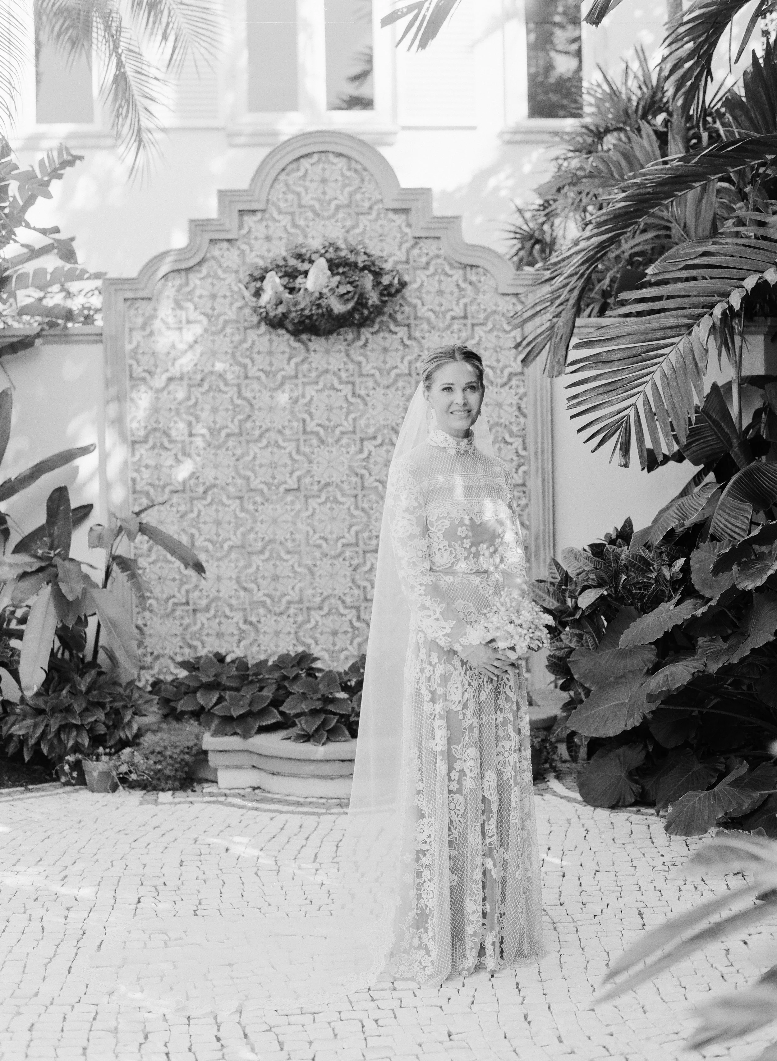 A bride in a lace wedding dress with a veil, standing outdoors in a garden with lush plants and a decorative backdrop.