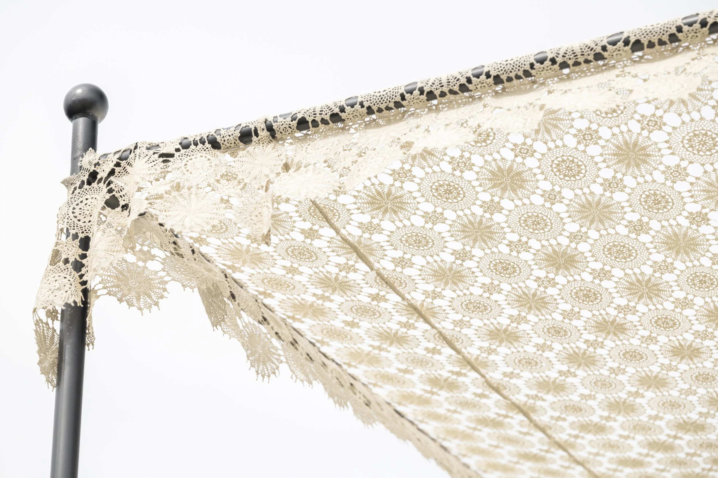 Close-up of an outdoor lace canopy with intricate floral and geometric patterns, supported by a black metal pole against a white sky.