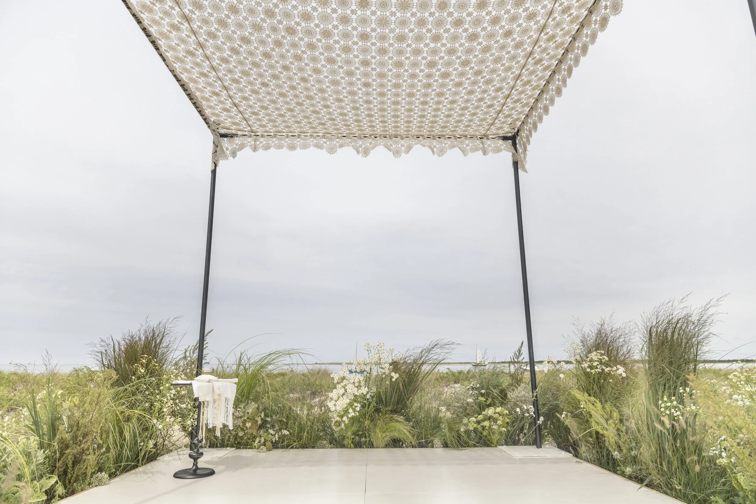 Outdoor wedding arch with black metal poles and a lace canopy, decorated with small white flowers, on a concrete platform with greenery and flowers in the background, under a cloudy sky.