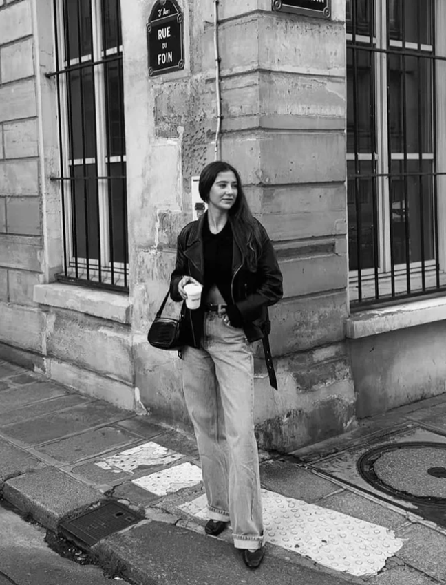 A young woman with long dark hair, wearing a black leather jacket, black top, light-colored jeans, and heels, standing on a city sidewalk in Paris. She is holding a coffee cup and a small purse, leaning against a corner of a building with iron-barred windows.