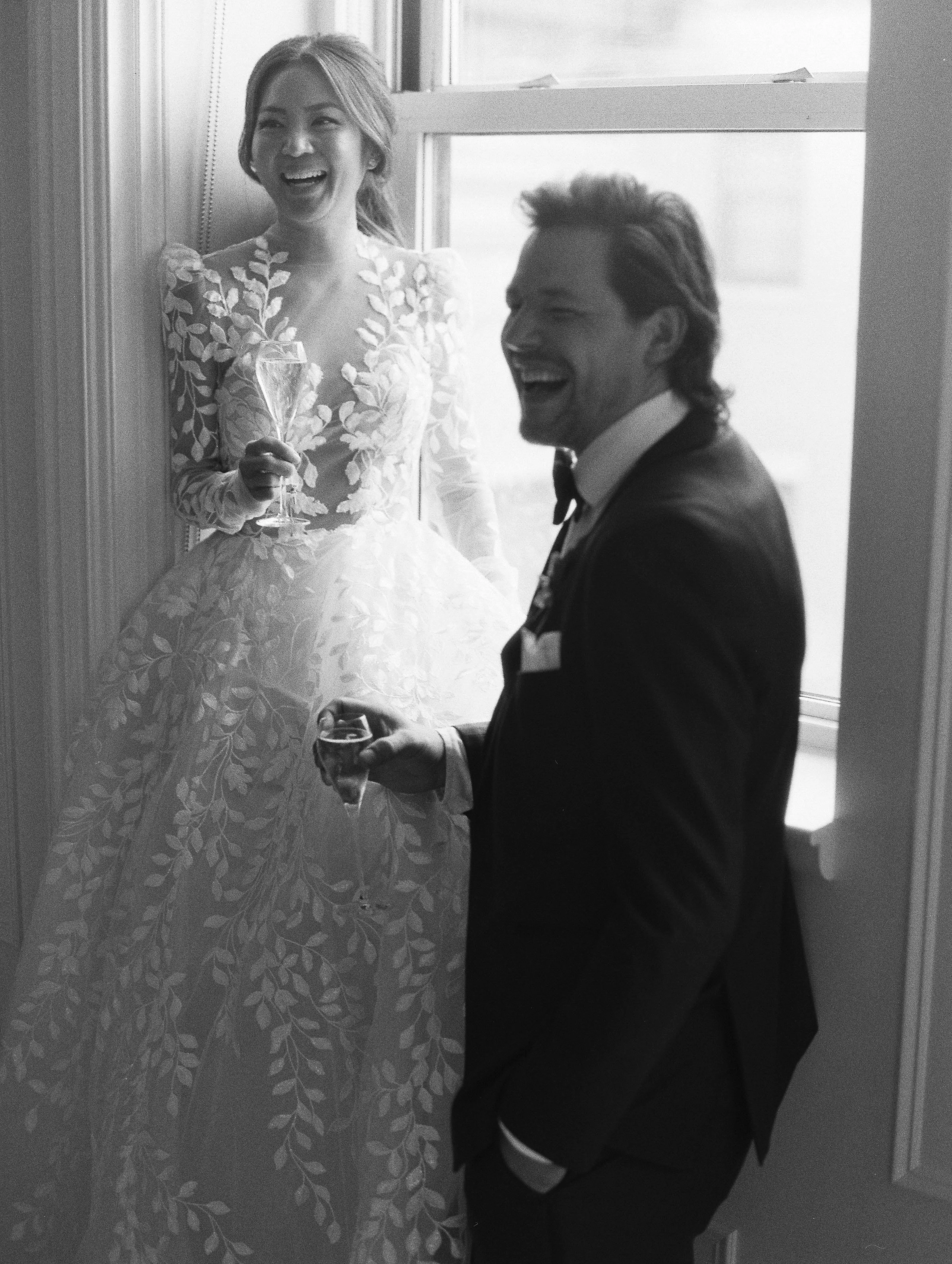 A black and white photo of a bride and groom celebrating with drinks, both laughing happily, in front of a window.