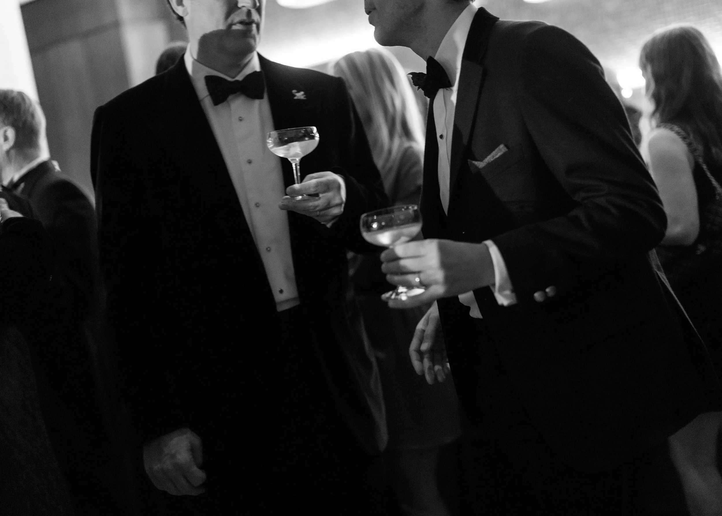 Two men in tuxedos holding cocktail glasses at a formal event, surrounded by other attendees.