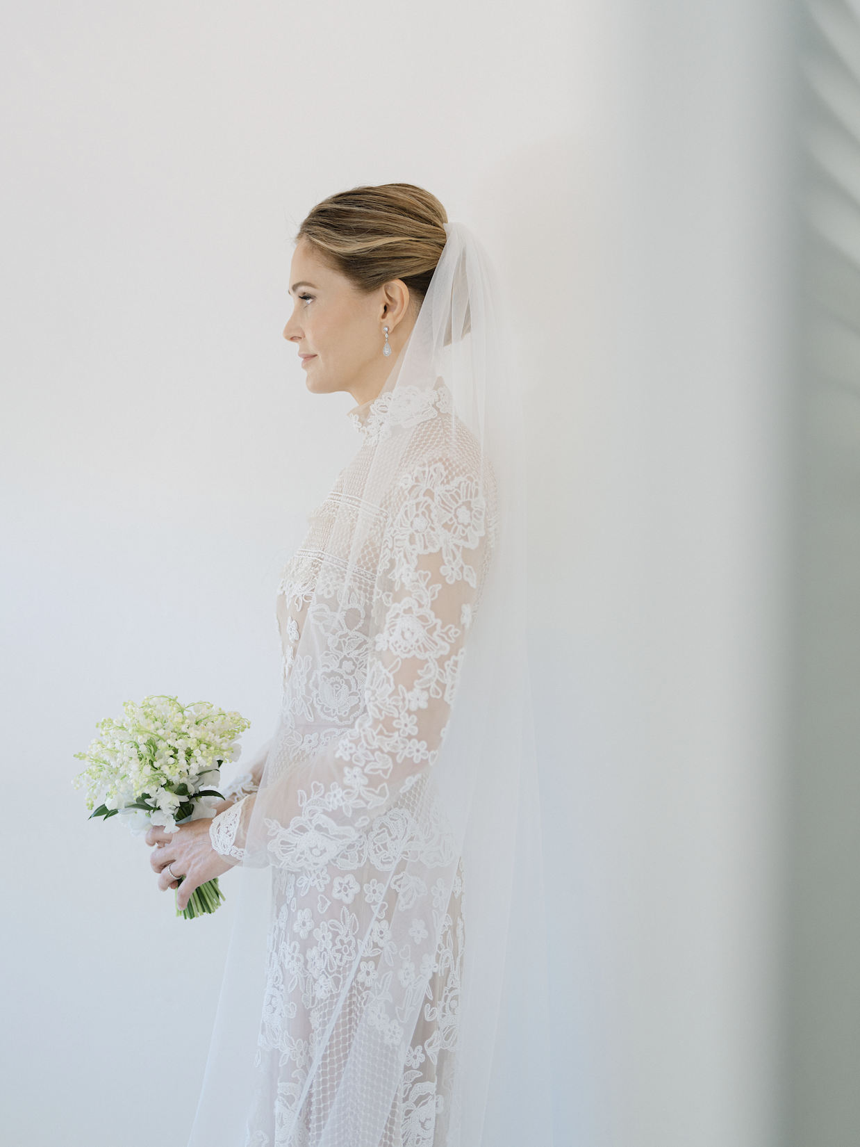 Brunette bride in lace wedding dress with lace high collar holding a small bouquet of white and green flowers, with a white veil flowing behind her, standing against a plain white background.