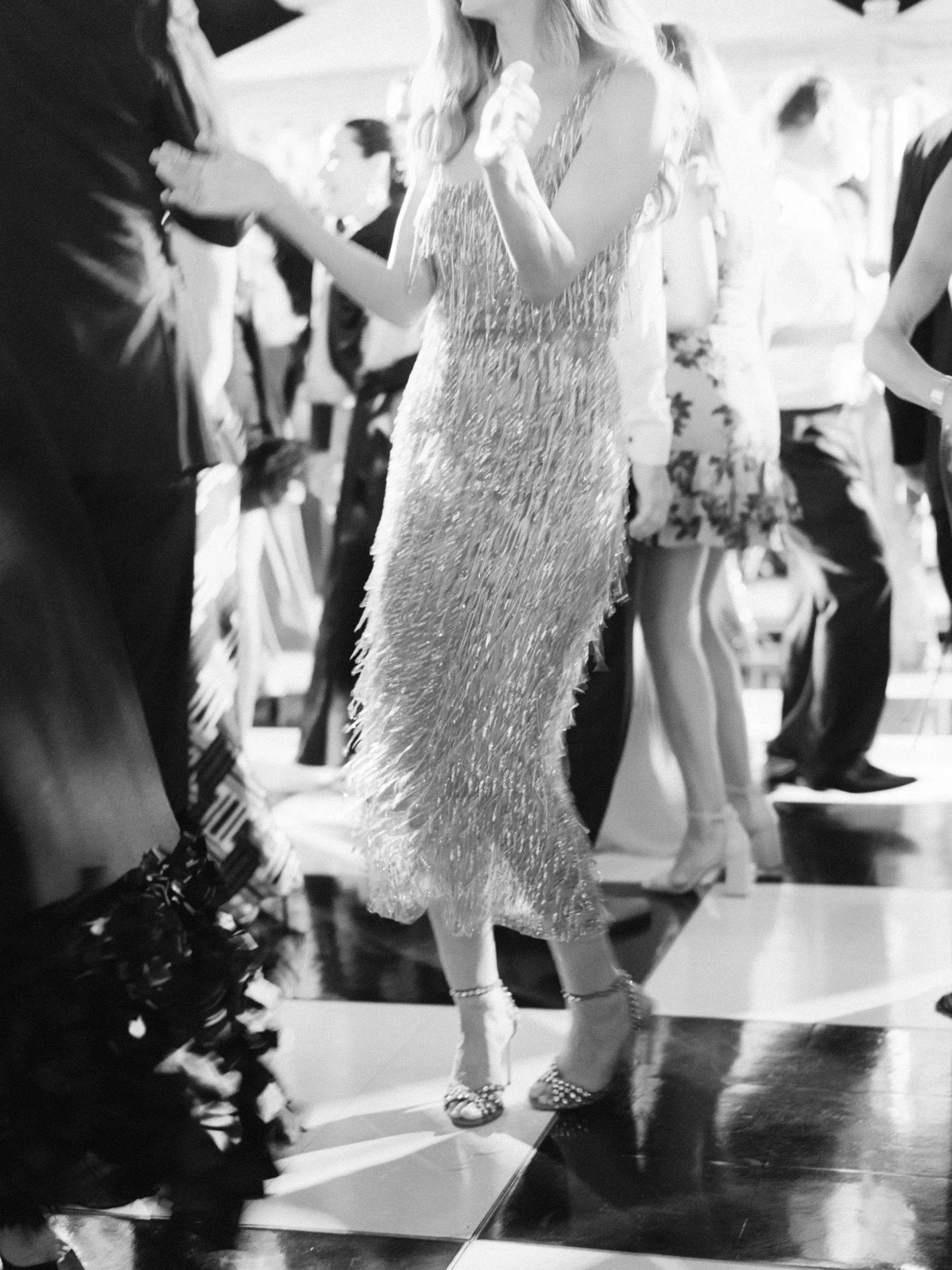 A woman in a shiny, long dress dancing with a man at a social event, with other people in the background.