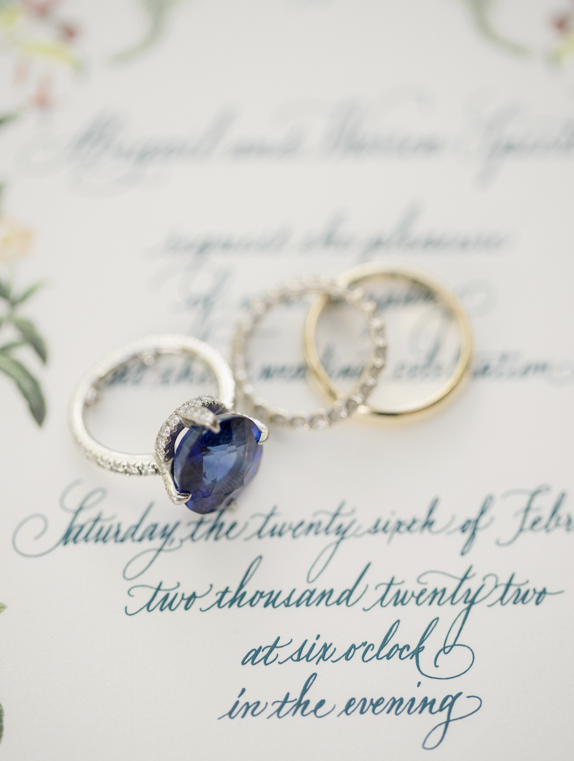 Close-up view of three rings placed on a wedding invitation with elegant cursive writing. One ring has a large dark blue gemstone surrounded by small diamonds, another is a simple gold band, and the third is a silver band with small diamonds.