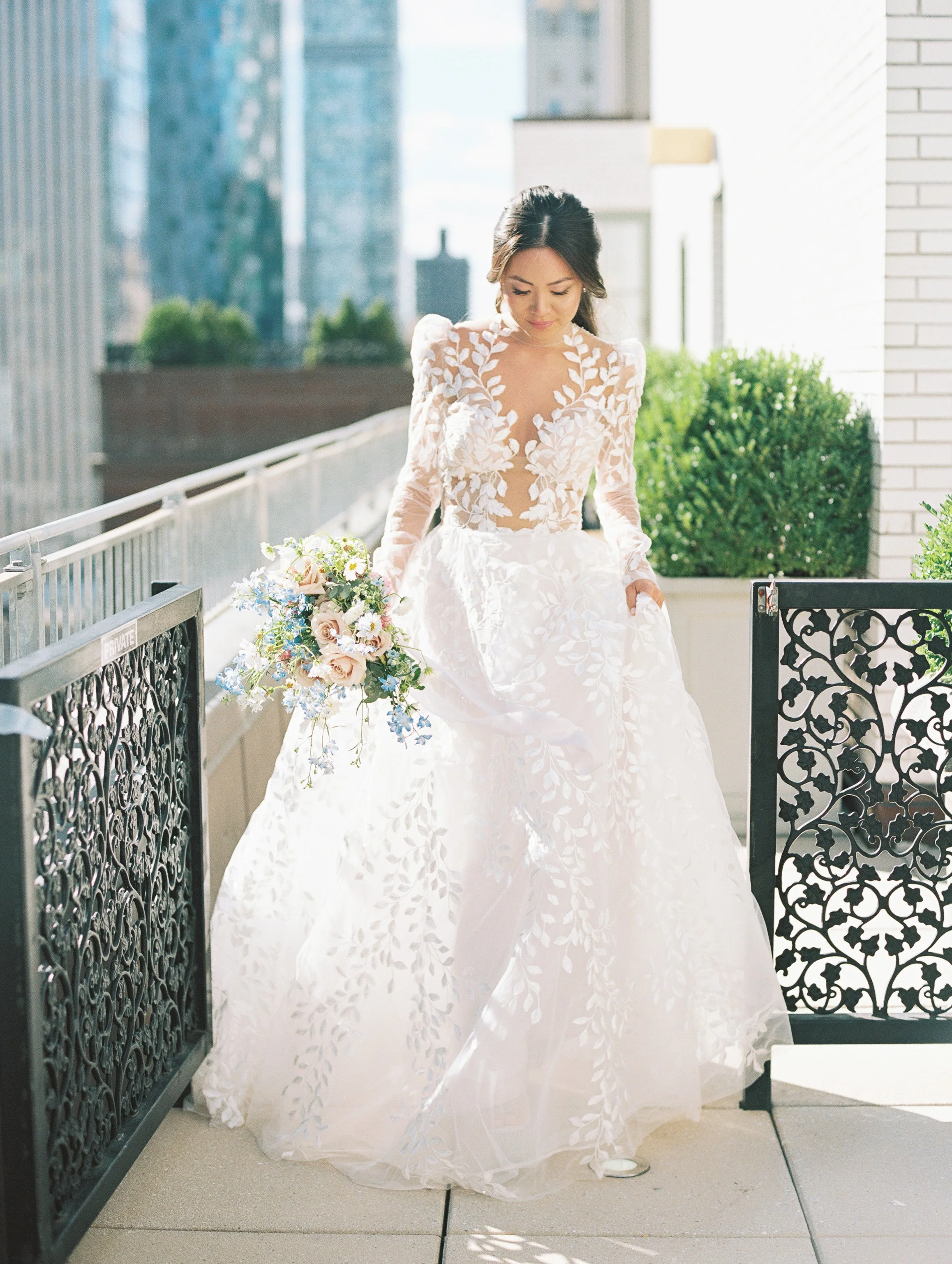 A bride in a white gown with floral lace details, holding a bouquet, walking outdoors on a balcony with city buildings in the background.