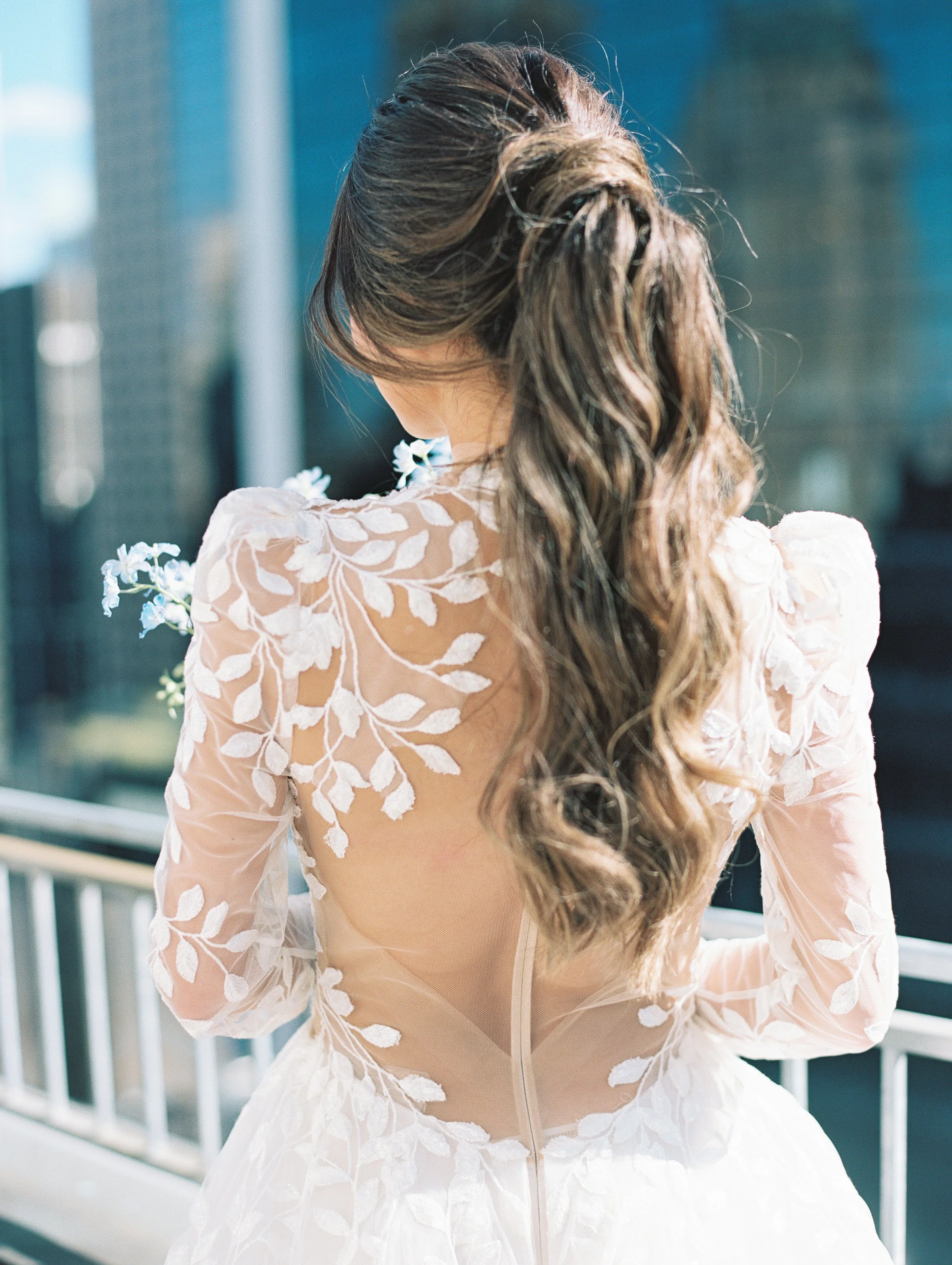 A woman with long, wavy brown hair wearing a white dress with sheer fabric and embroidered leaf patterns, standing on a balcony with an urban cityscape background.