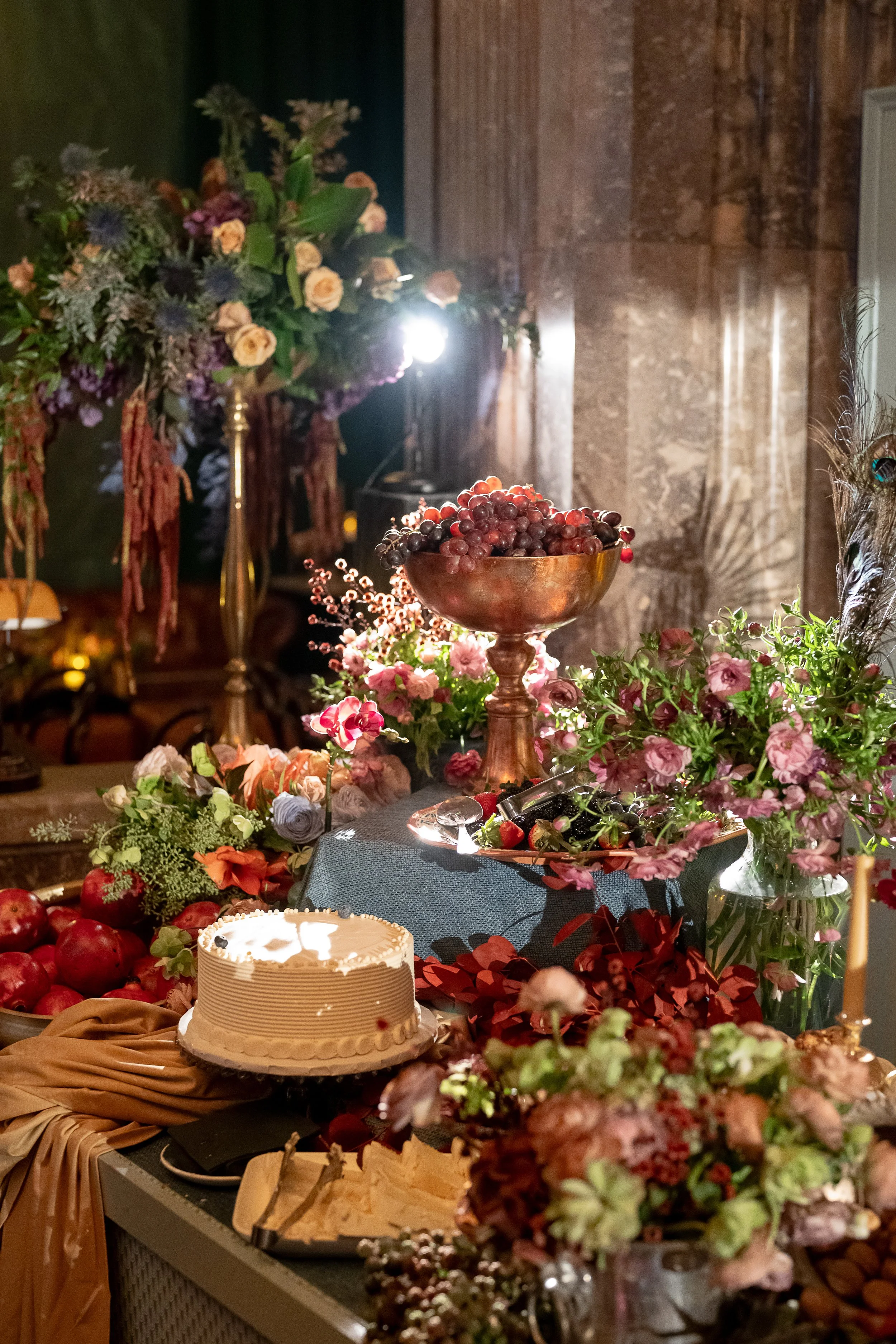 A table decorated with flowers, fruits, and a cake, with a large bowl of grapes in the center and soft warm lighting.