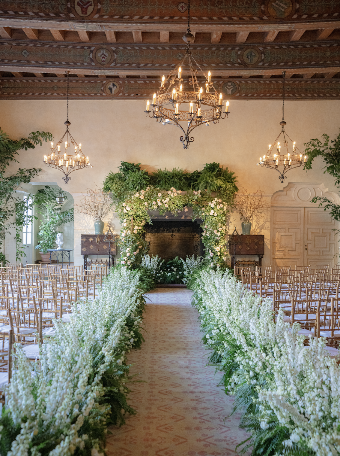 Elegant indoor wedding ceremony space with a floral arch and aisle, surrounded by rows of wooden chairs, chandeliers hanging from a decorated ceiling, green plants, and natural light.