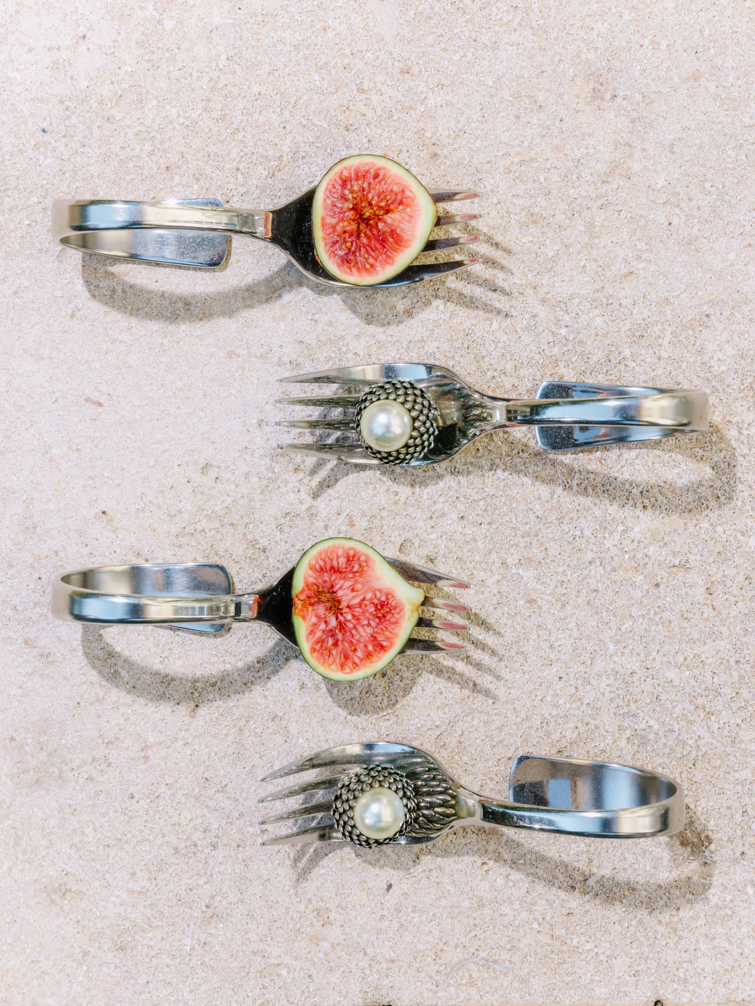 Four metal spoons with decorative handles, two of which hold halved figs and two hold jewelry brooches with pearl and floral details, arranged on a textured surface.