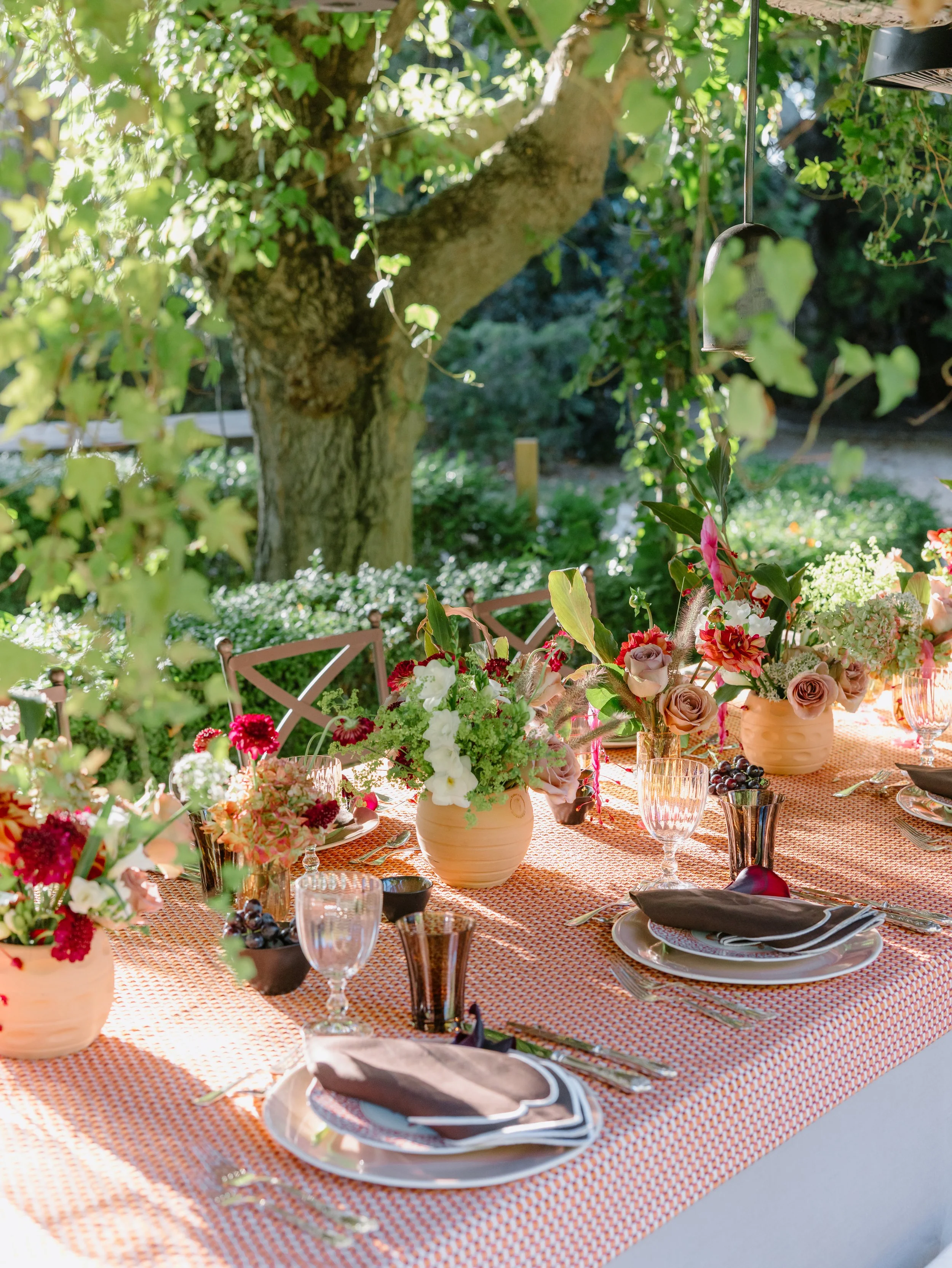 A table set for outdoor dining with floral arrangements, glassware, plates, and cutlery, beside a large tree and lush greenery.