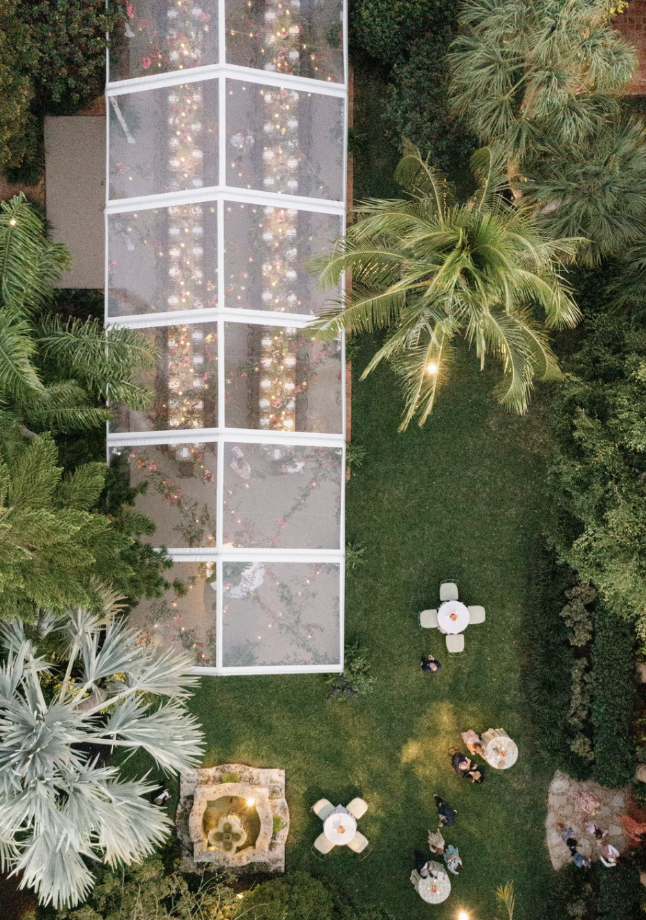 Aerial view of an outdoor garden with a large rectangular structure with glass panels and string lights inside, surrounded by lush green trees and several tables with chairs, some with white tablecloths and decorations.