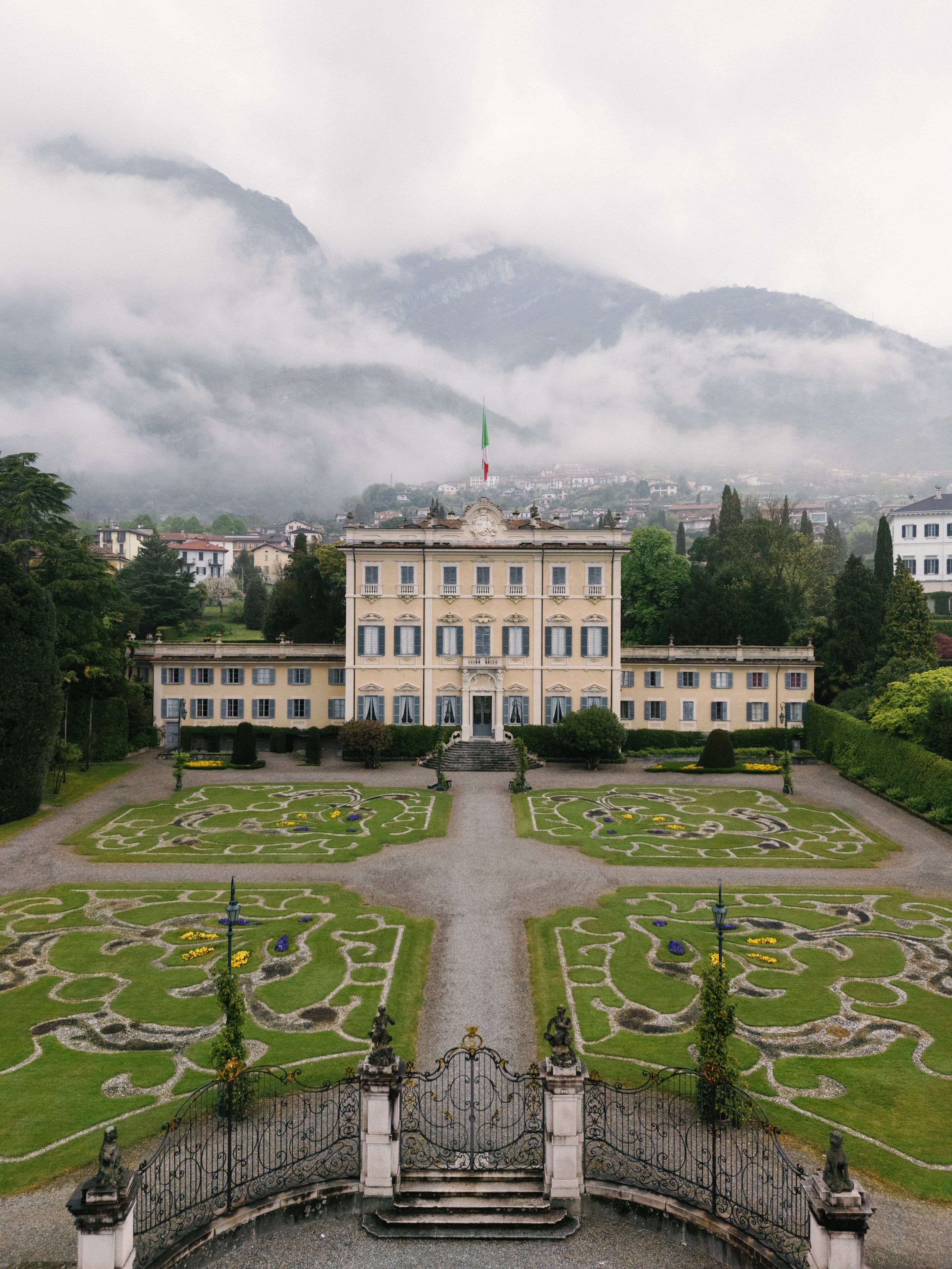 A large yellow mansion with blue shutters, surrounded by a manicured garden with symmetrical flower beds and a wrought iron gate, set against foggy mountains in the background.