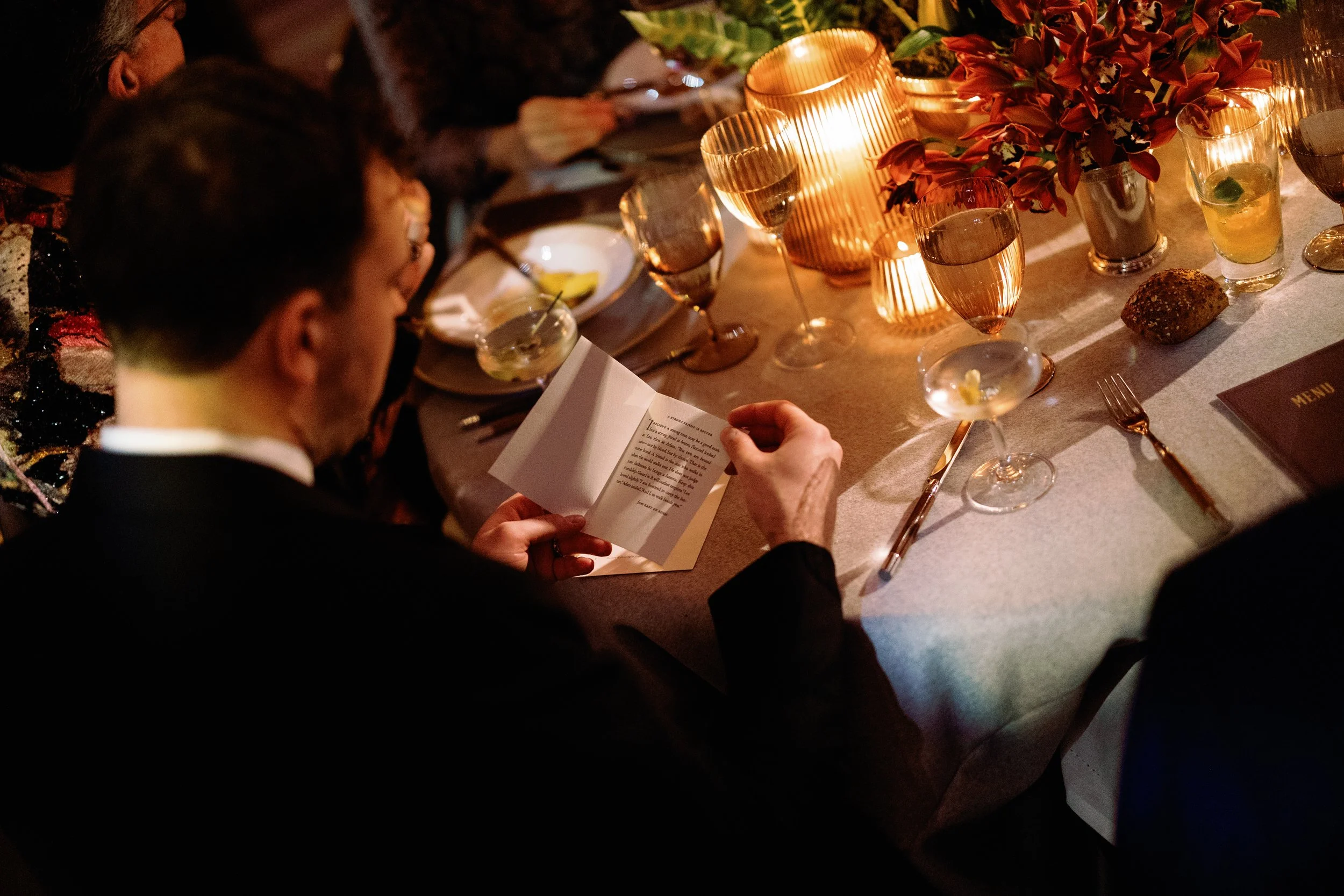 A person in a dark suit reading a card at a dinner table decorated with flowers and lit candles, surrounded by glassware, plates, and a menu.