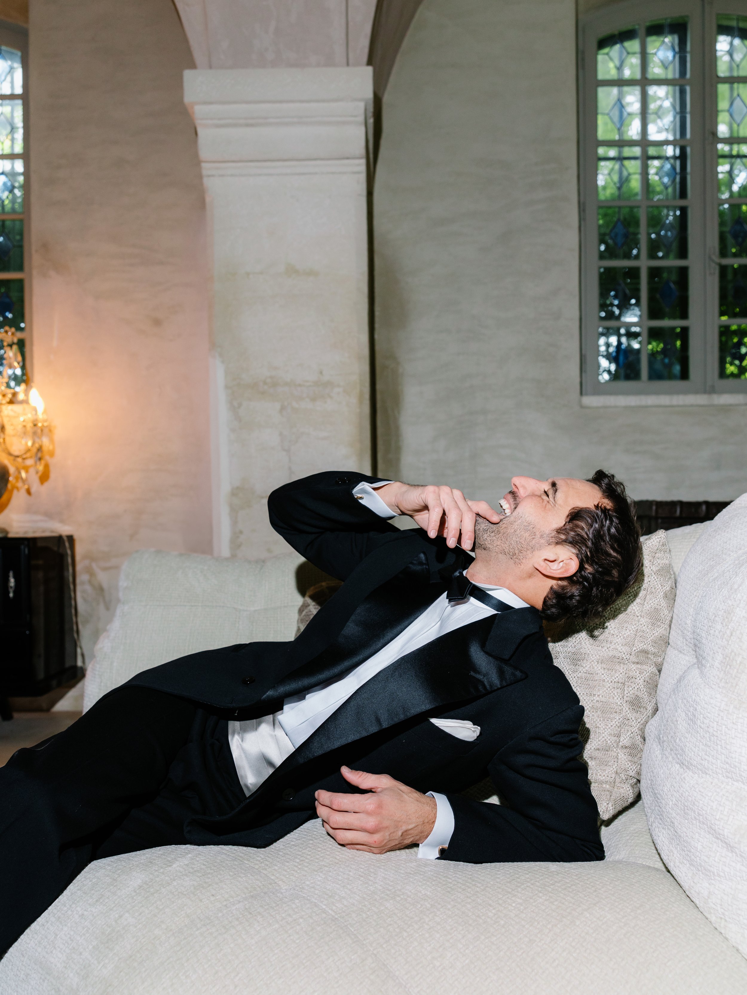 Man in tuxedo laughing and lying on a couch in a room with large windows.