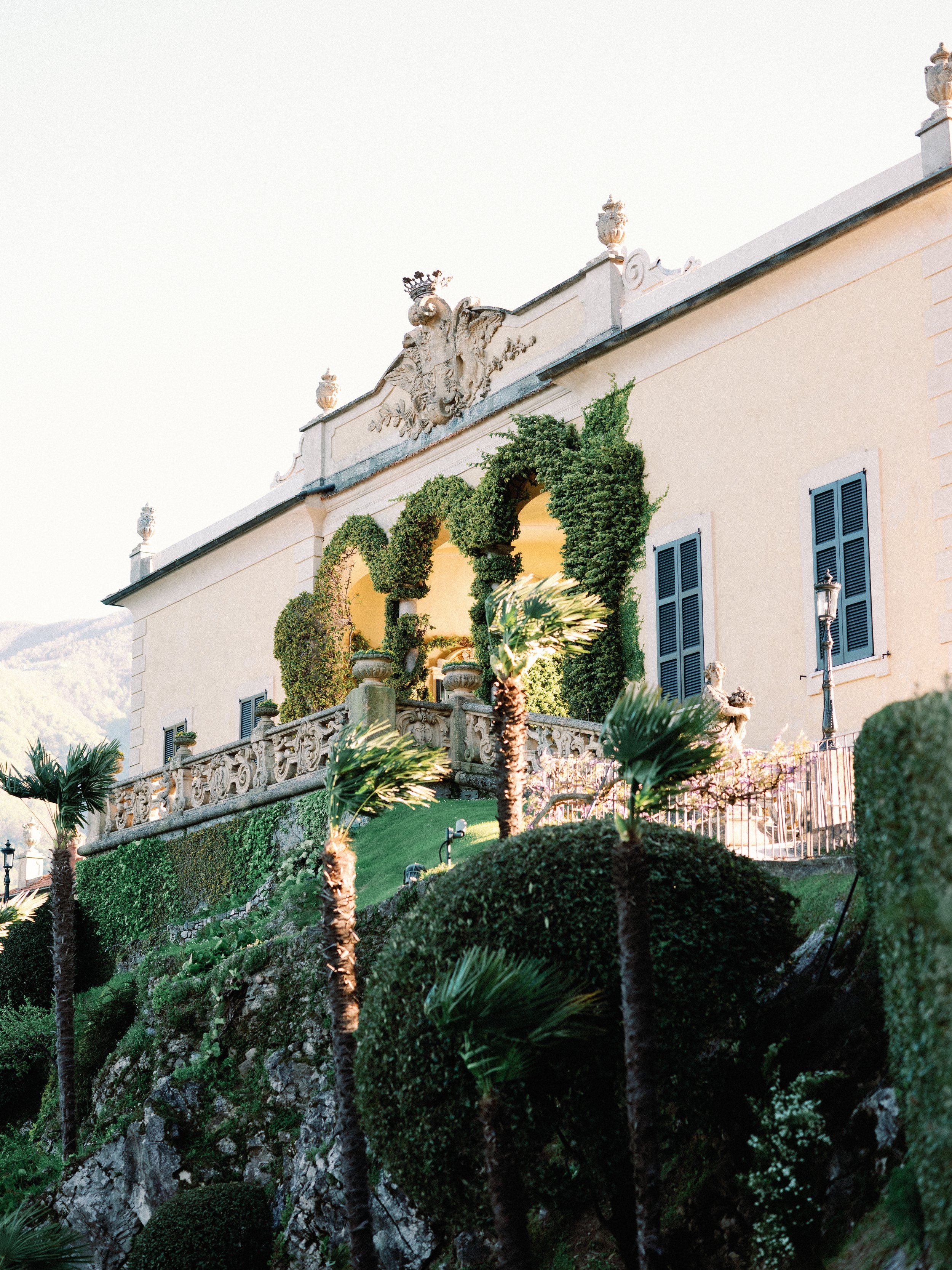 Elegant mansion with ornate crest on roof, surrounded by lush greenery, palms, and decorative wrought iron fencing.
