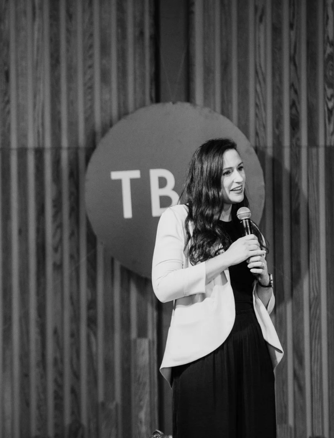 A woman with long dark hair, wearing a white blazer and black dress, speaking into a microphone on stage in front of a wooden wall and a circular sign with the letters 'TB' in a comedy or presentation setting.