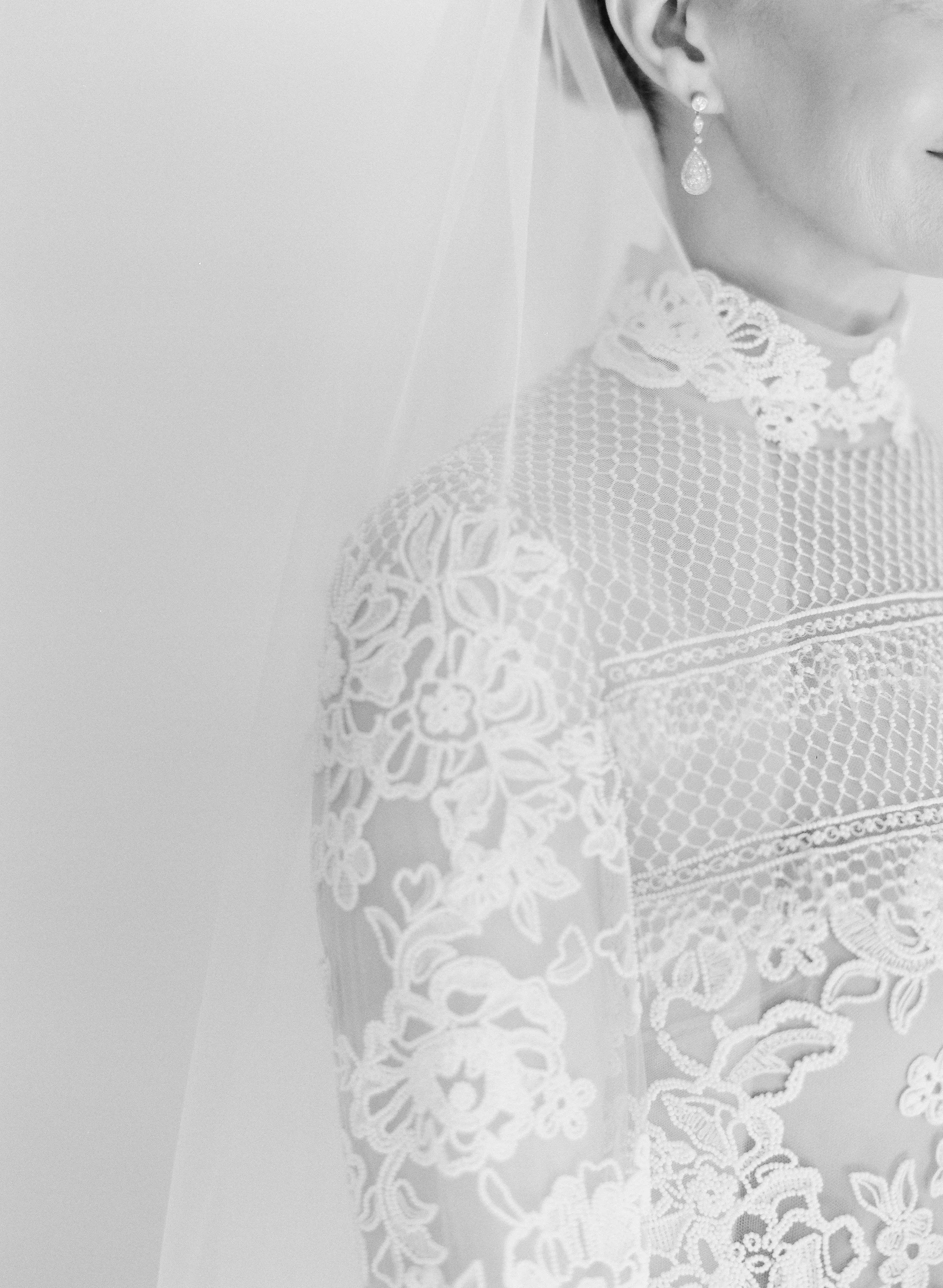 Close-up of a bride wearing a lace wedding dress with floral embroidery, pearl earrings, and a veil.