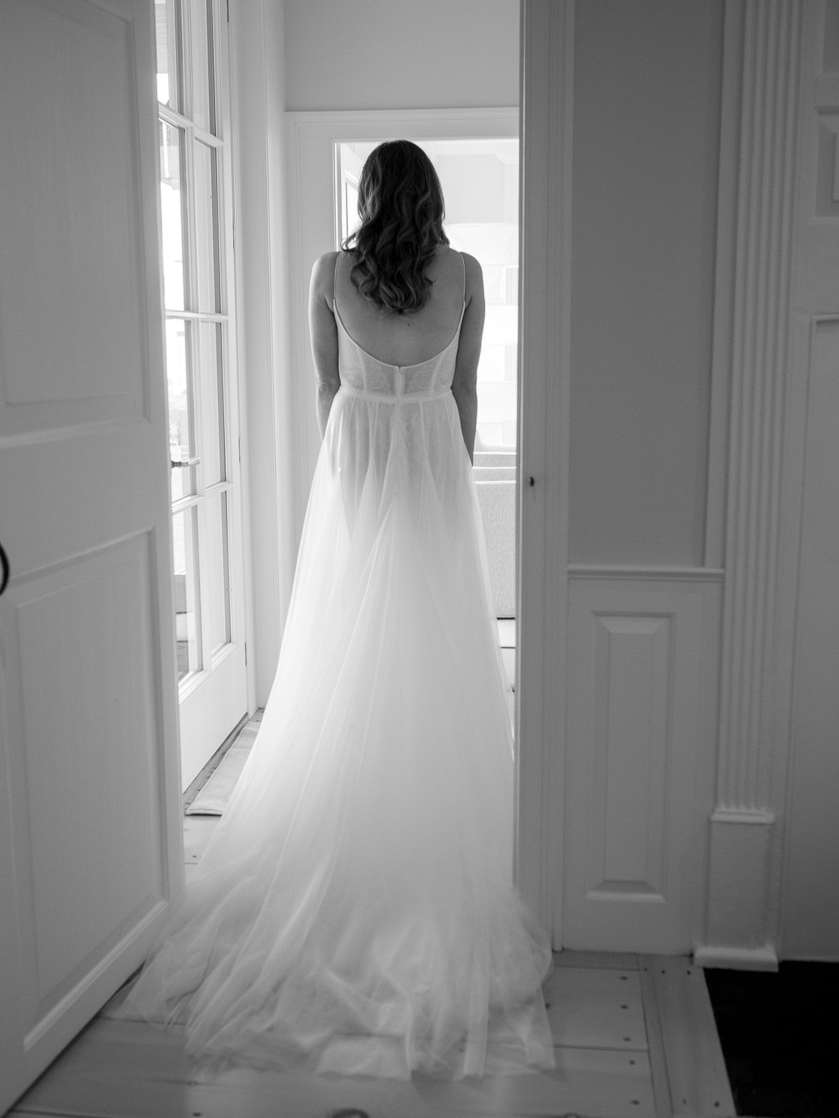 A woman in a wedding dress standing in a doorway, facing away, looking out through a window.