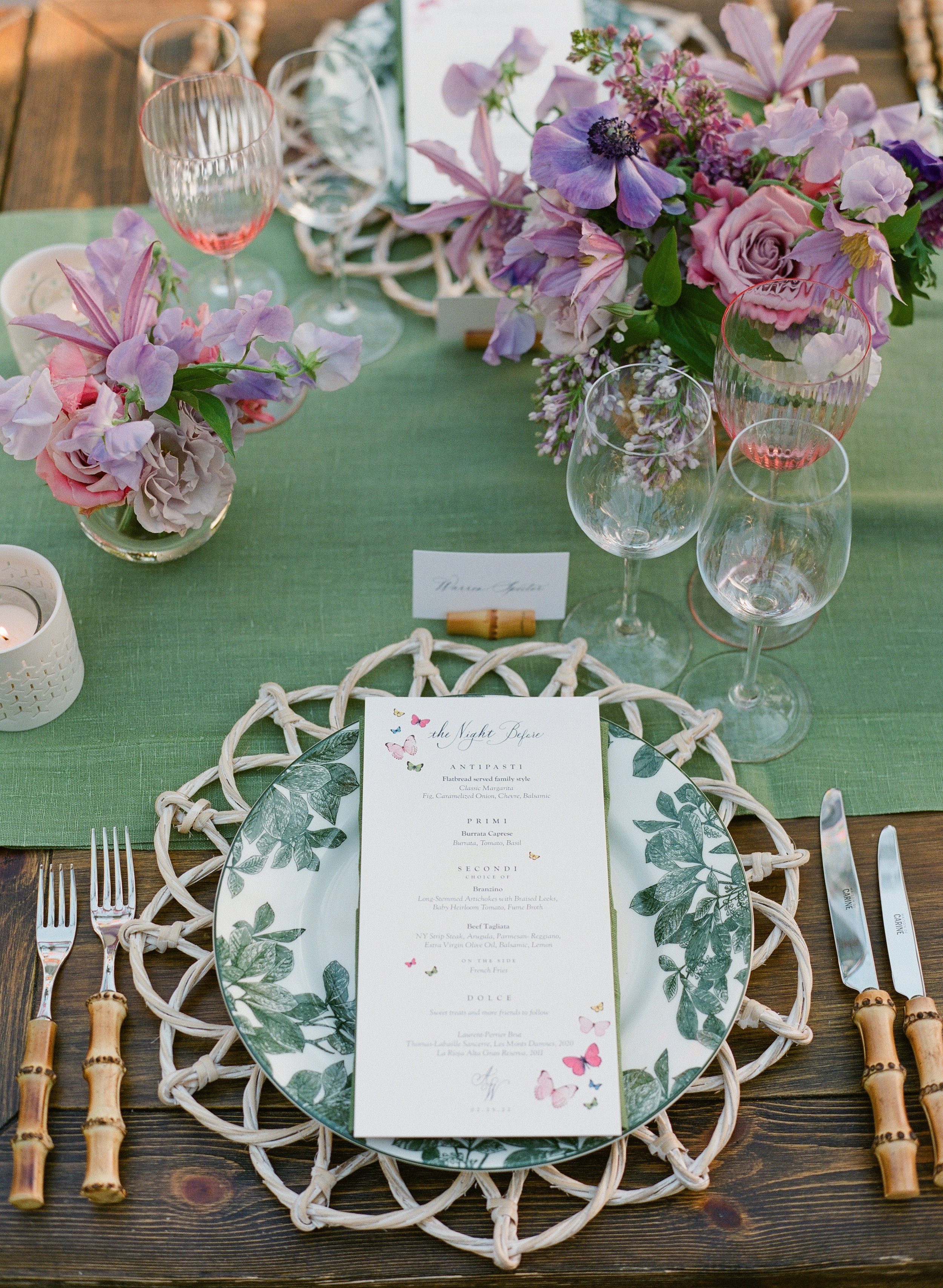 A beautifully set dining table featuring floral centerpieces, empty wine glasses, a candle holder, and a detailed menu card on a decorative plate. The table is decorated with a green table runner and bamboo-style cutlery.