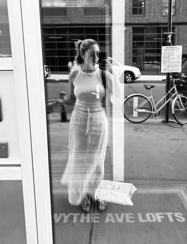 A woman taking a mirror selfie on an urban sidewalk, wearing a tank top, a long skirt, and sunglasses, with parked bicycles and cars visible outside.