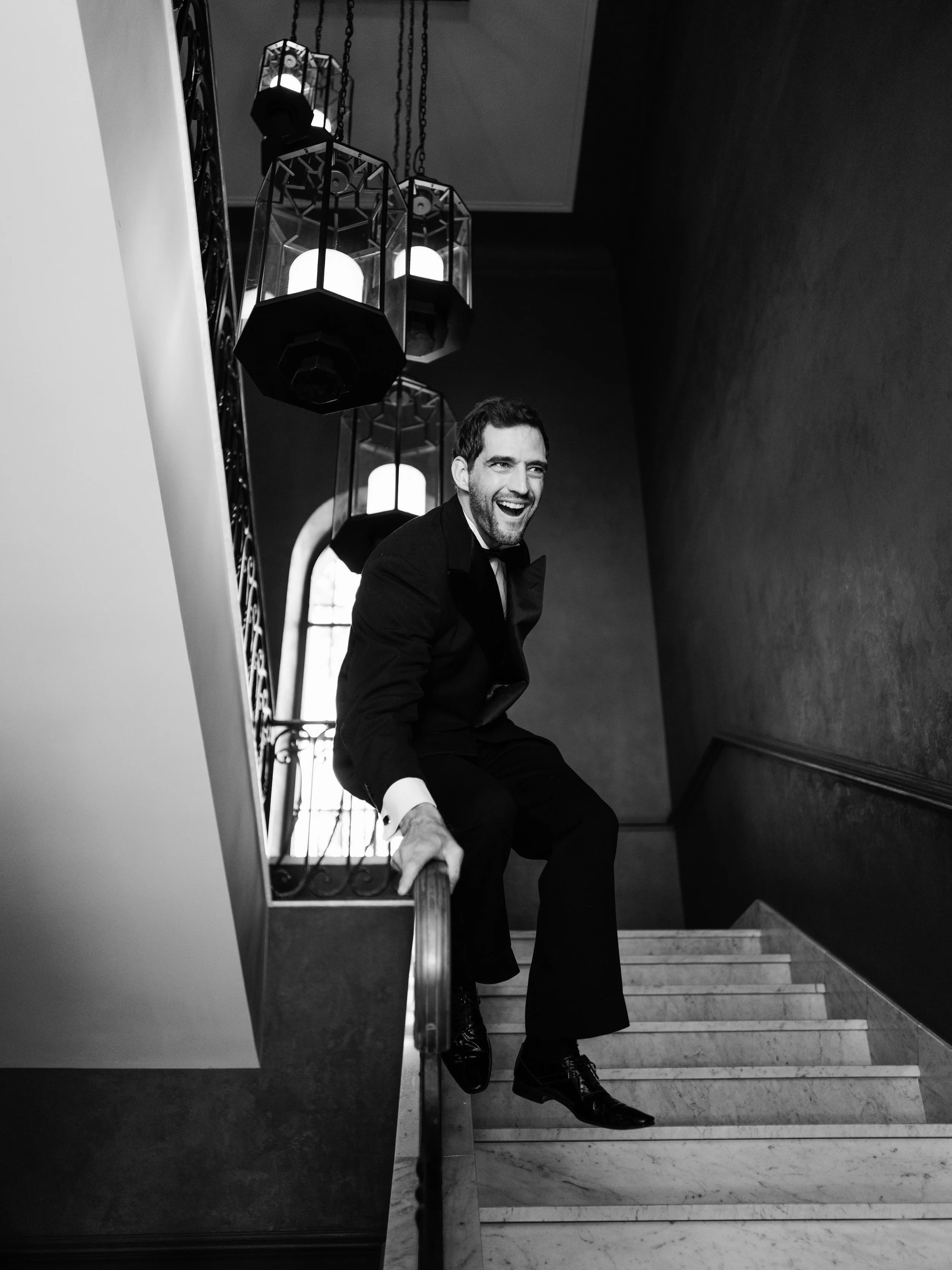 A man in formal attire, sitting on a staircase, smiling, with a decorative railing and hanging lights in the background.