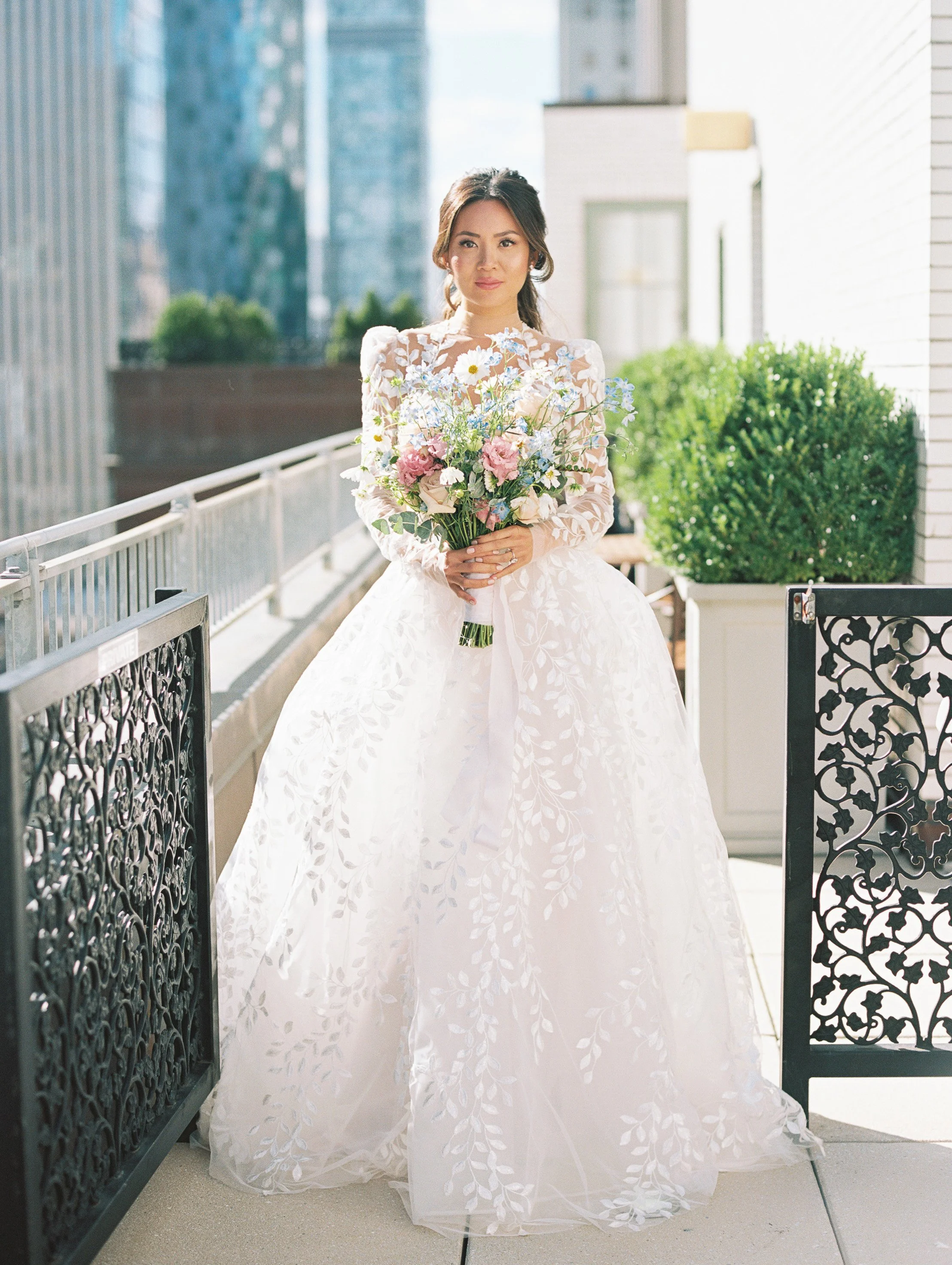 A woman in a white wedding dress holding a bouquet of flowers on a balcony with a cityscape in the background.