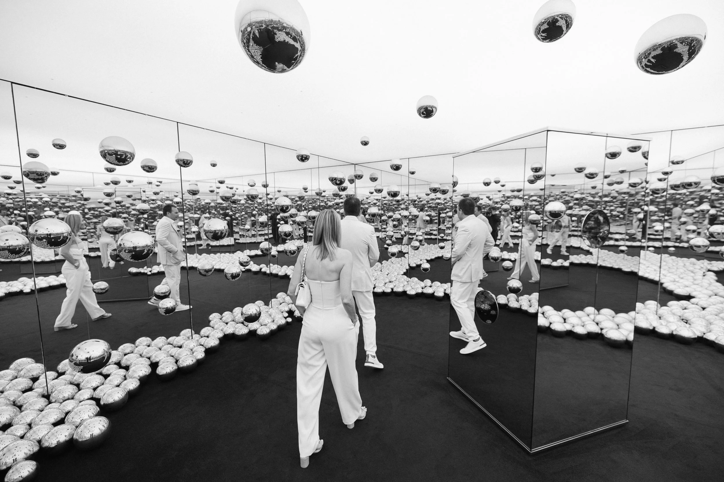 People inside a mirrored room installation with numerous reflective spheres on the floor and hanging from the ceiling, creating a disorienting, kaleidoscopic effect.