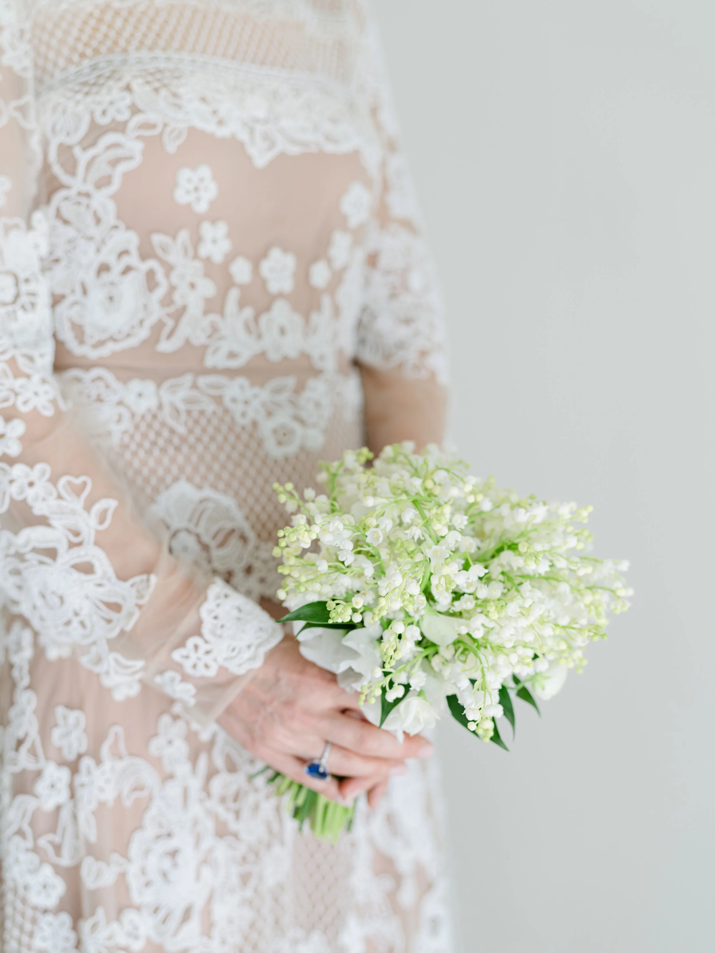 A woman in a lacy, sheer white dress holding a bouquet of white and green flowers.
