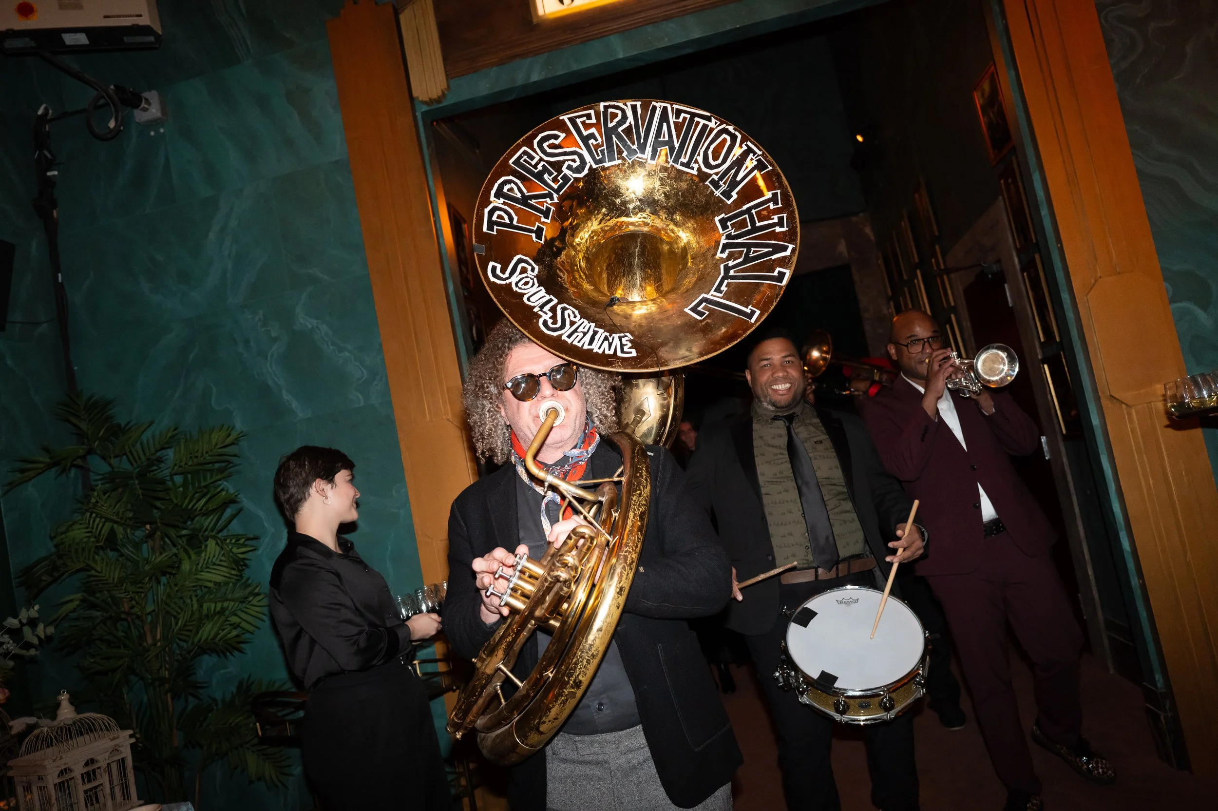 A jazz band performing at an event, with a man playing a sousaphone and others playing a drum and trumpet, inside a venue with green and wood-paneled walls.