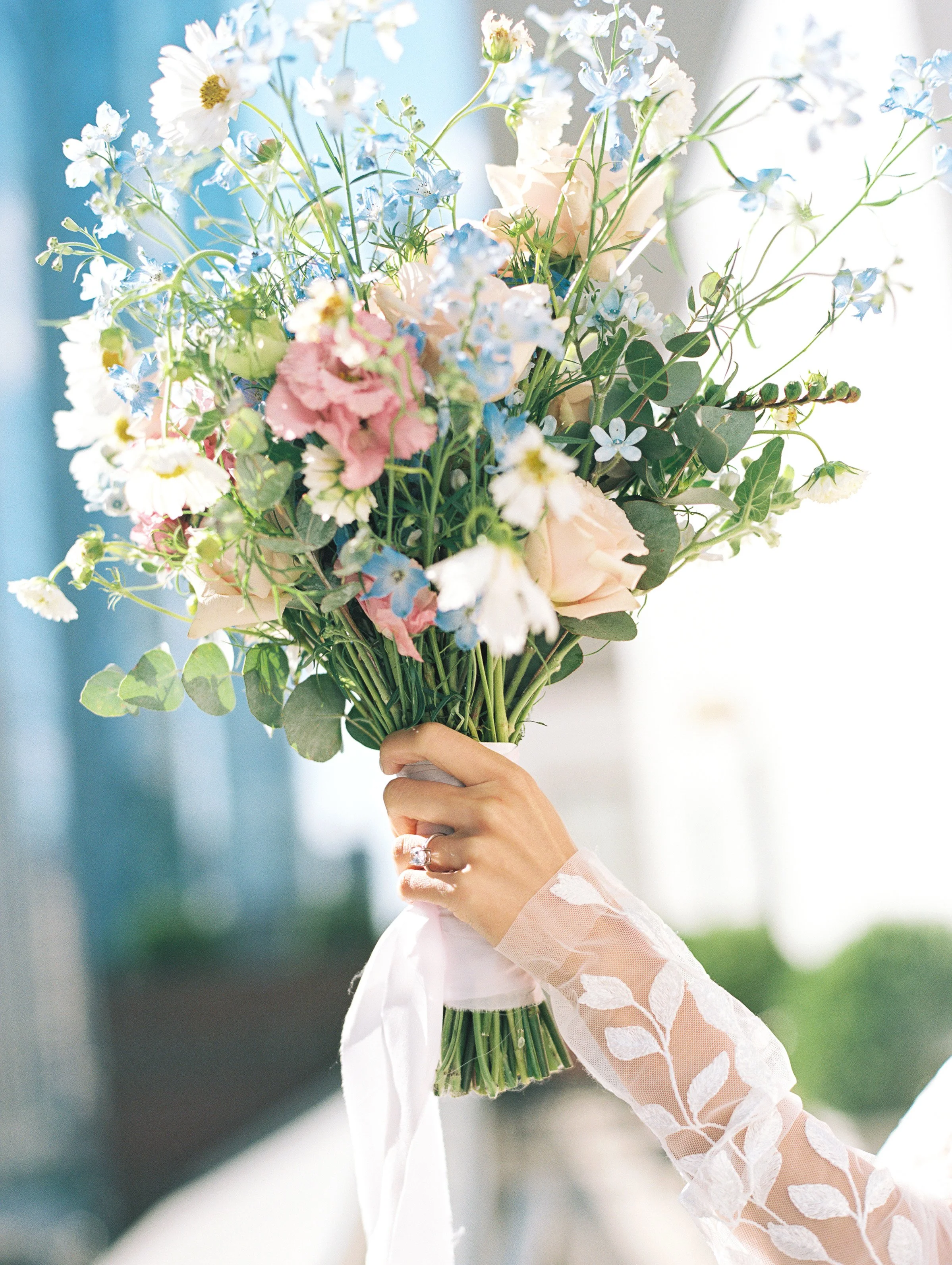 Person holding a bouquet of mixed flowers including roses and wildflowers with lace sleeve in sunlight.