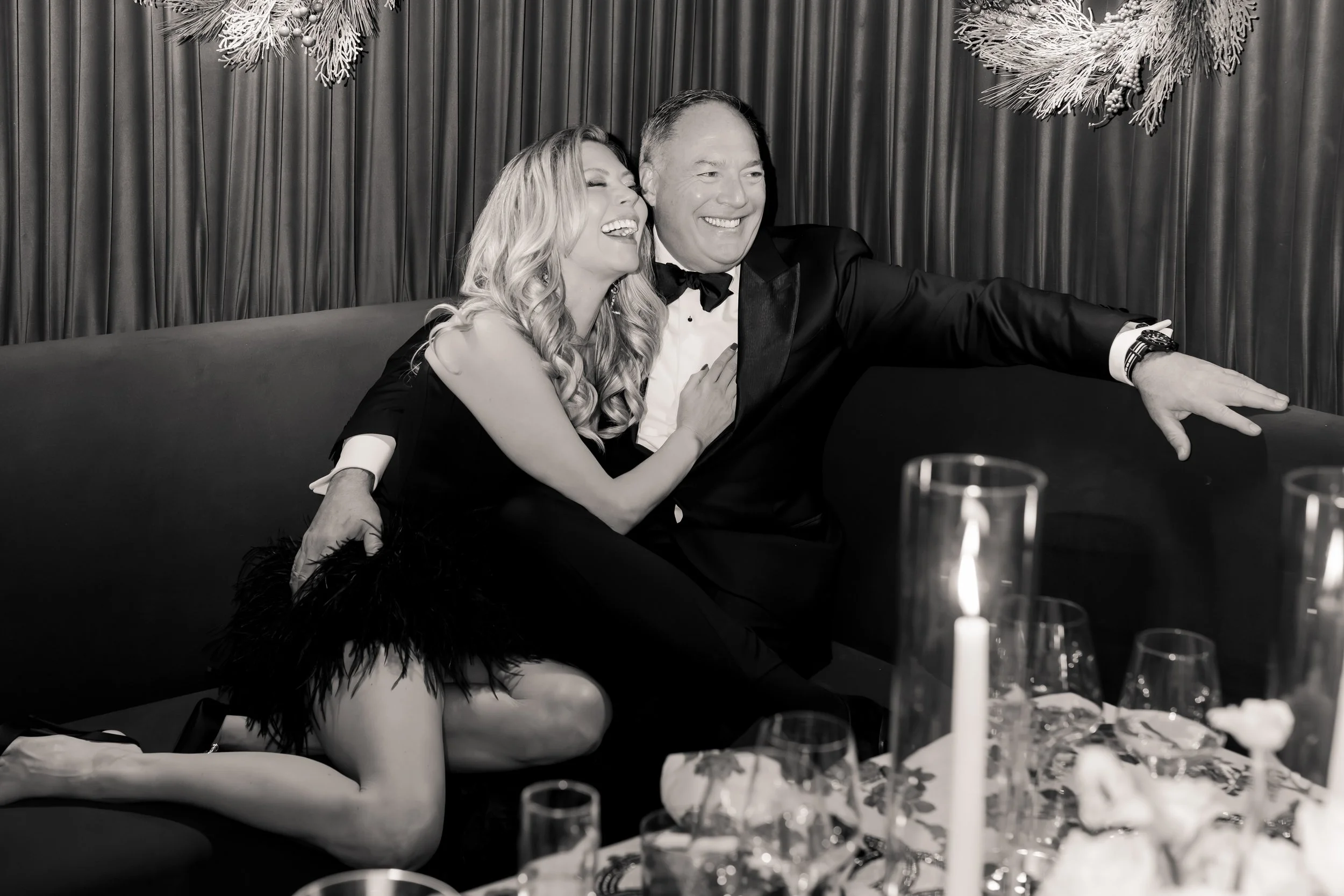 A man and woman dressed in formal attire sitting on a sofa, laughing and enjoying themselves at a celebration or party, with tableware and candles in the foreground.