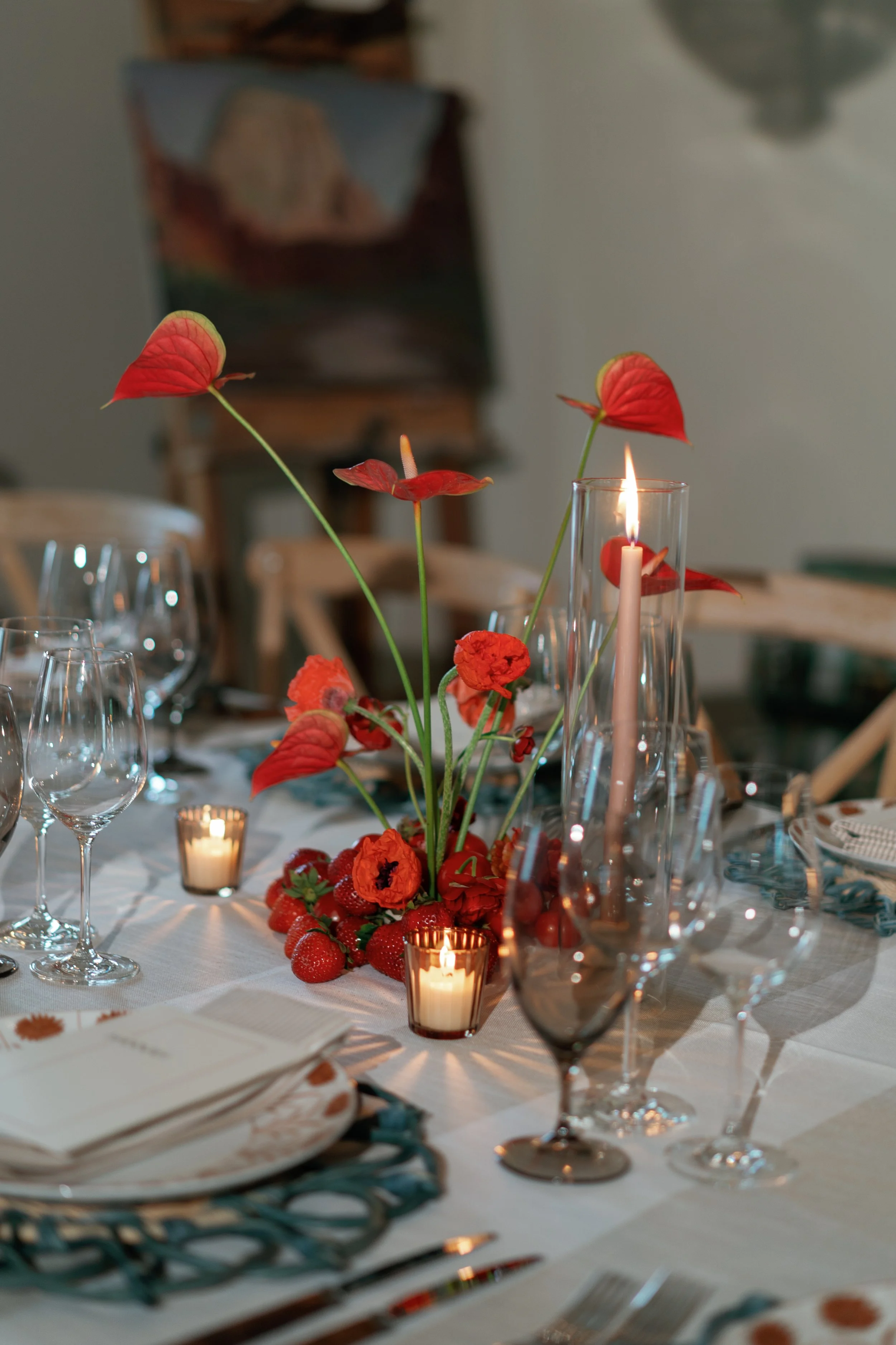 Decorated dining table featuring red flowers, strawberries, candles, and glassware, set for a formal meal.