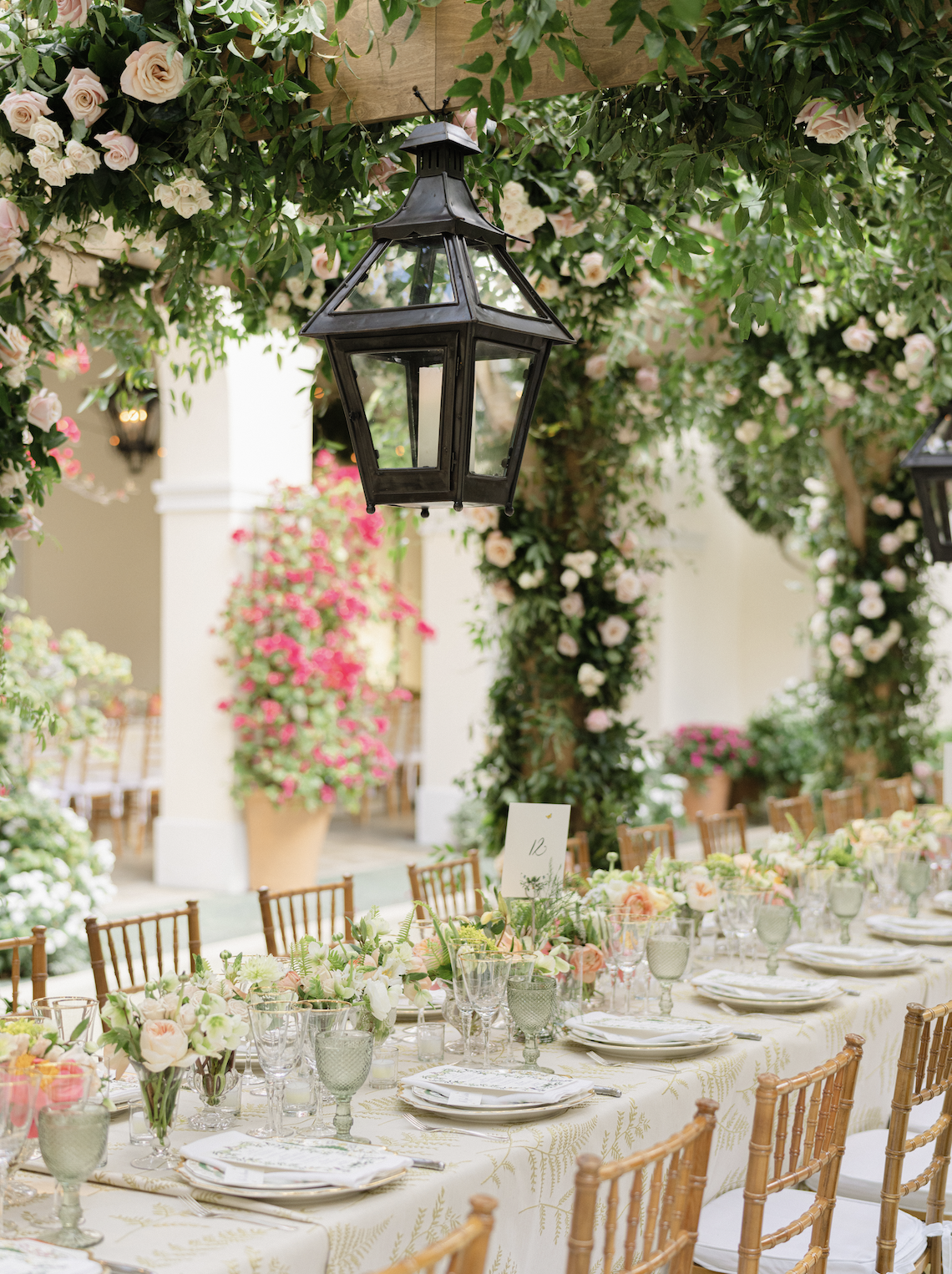 Elegant outdoor banquet table decorated with floral centerpieces, surrounded by wooden chairs, under hanging black lanterns, with lush greenery and flowers in the background.